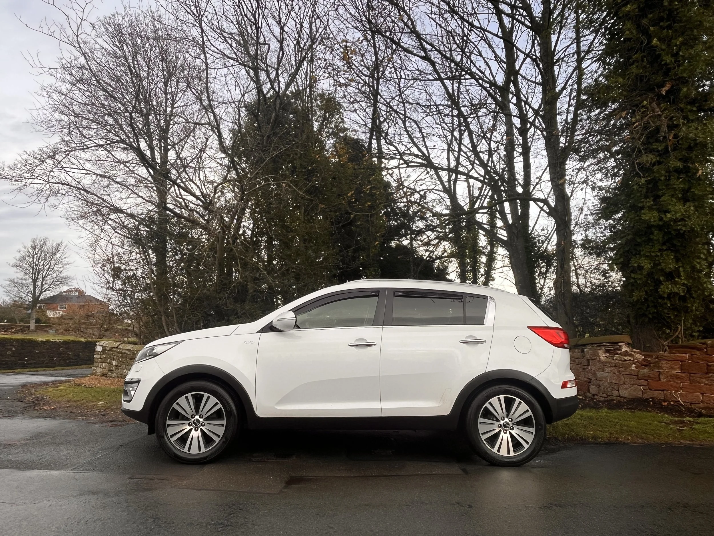 A white compact SUV parked on a wet road in front of a low stone wall, with leafless trees and a cloudy sky in the background.