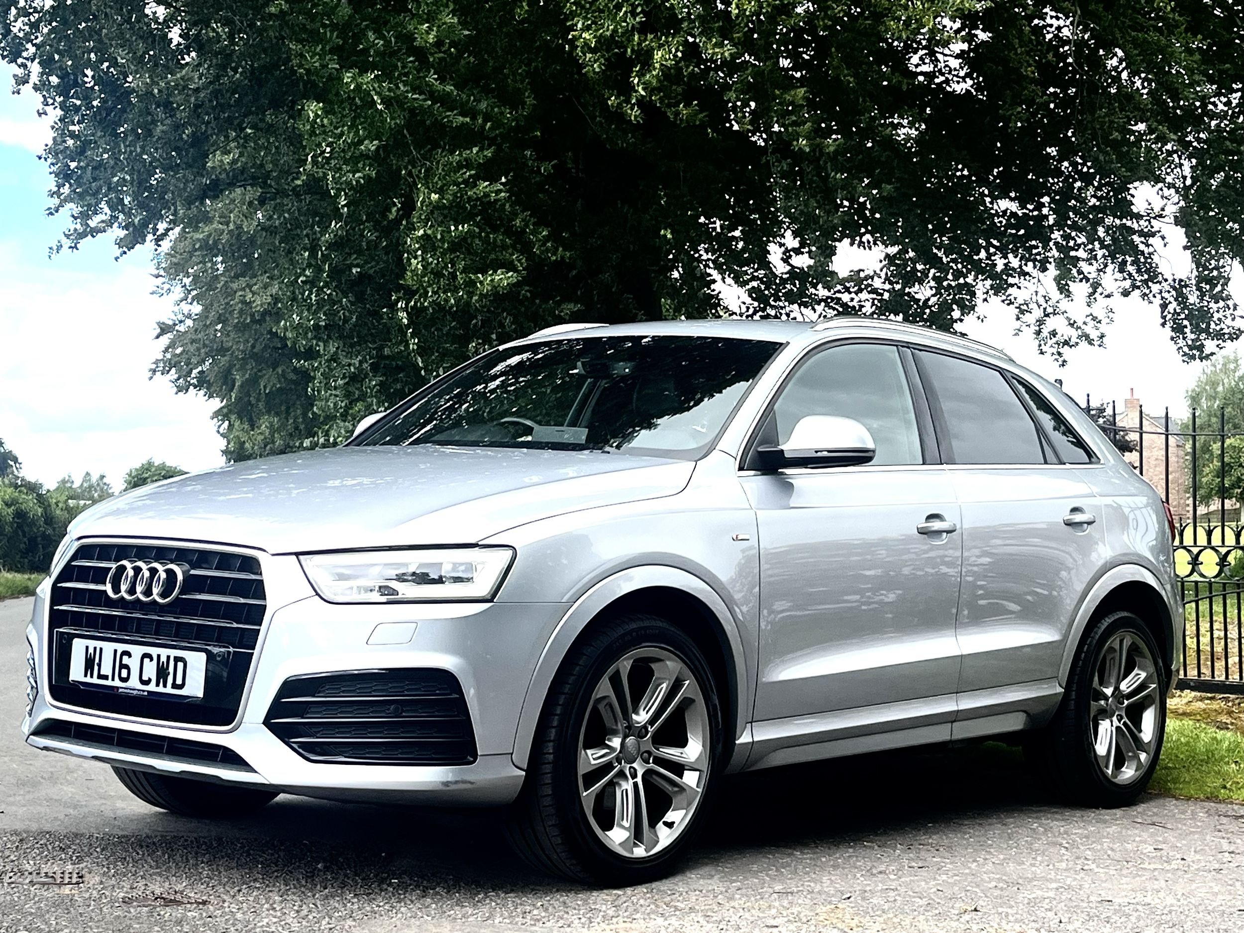 A silver Audi SUV parked outdoors under a large green tree, with a house and a black fence in the background.