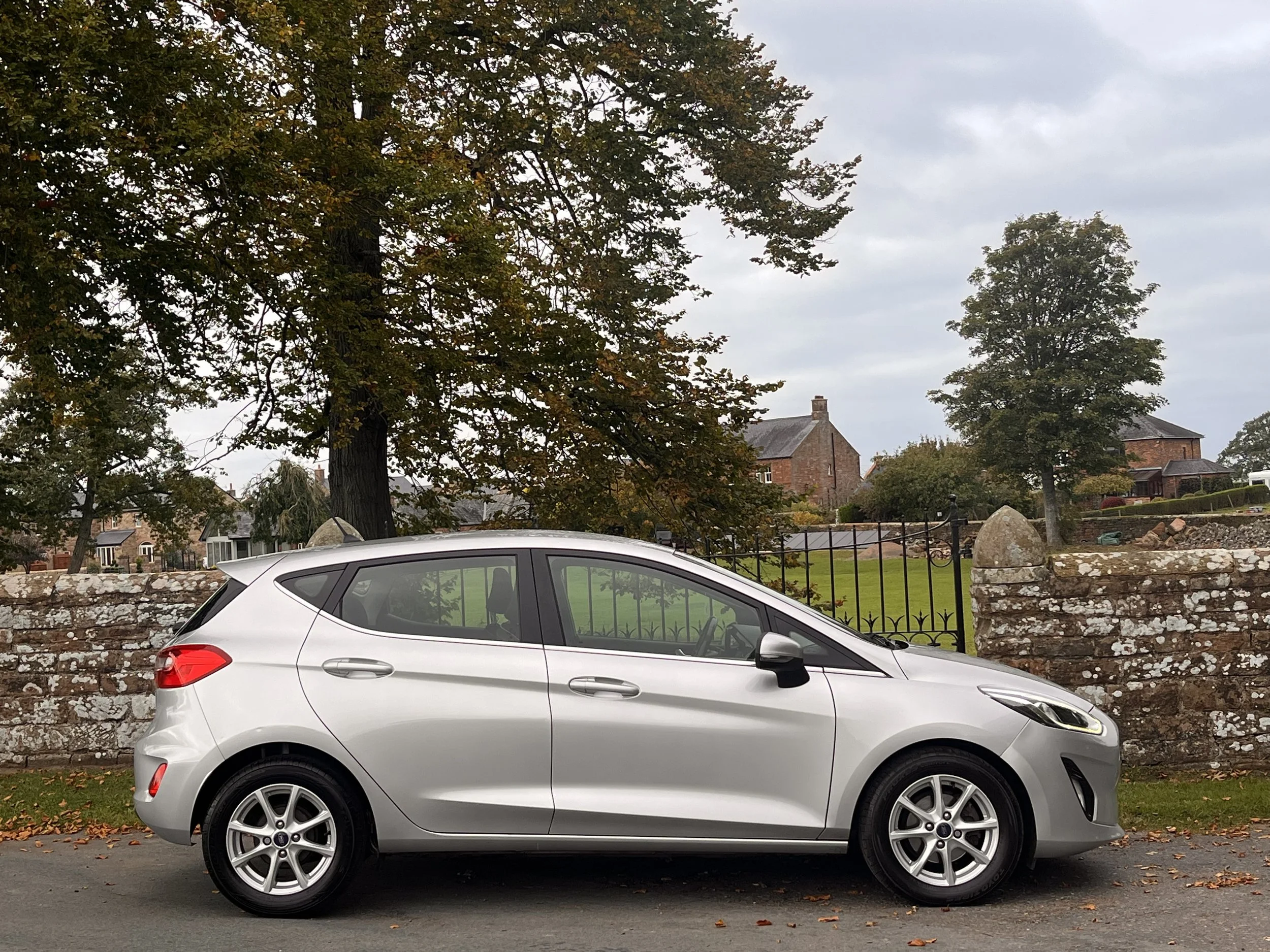 A silver hatchback car parked on the street in front of a stone wall and gate, with trees and houses in the background under a cloudy sky.