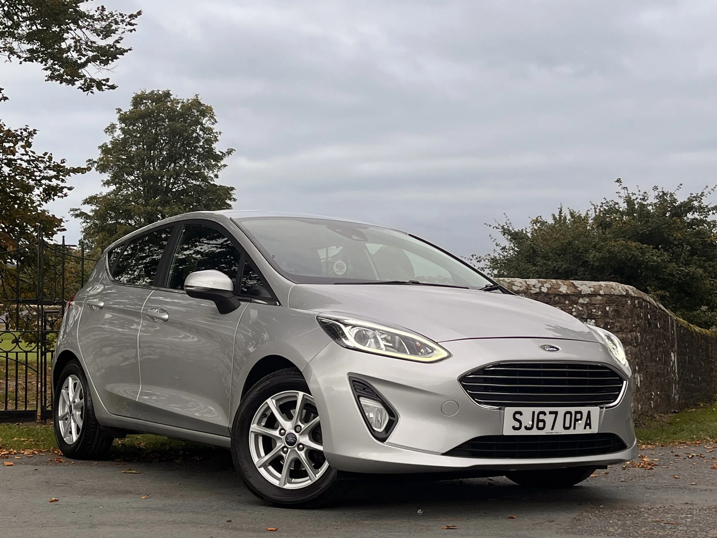 Silver Ford Fiesta parked on a street with trees and a stone wall in the background under a cloudy sky.