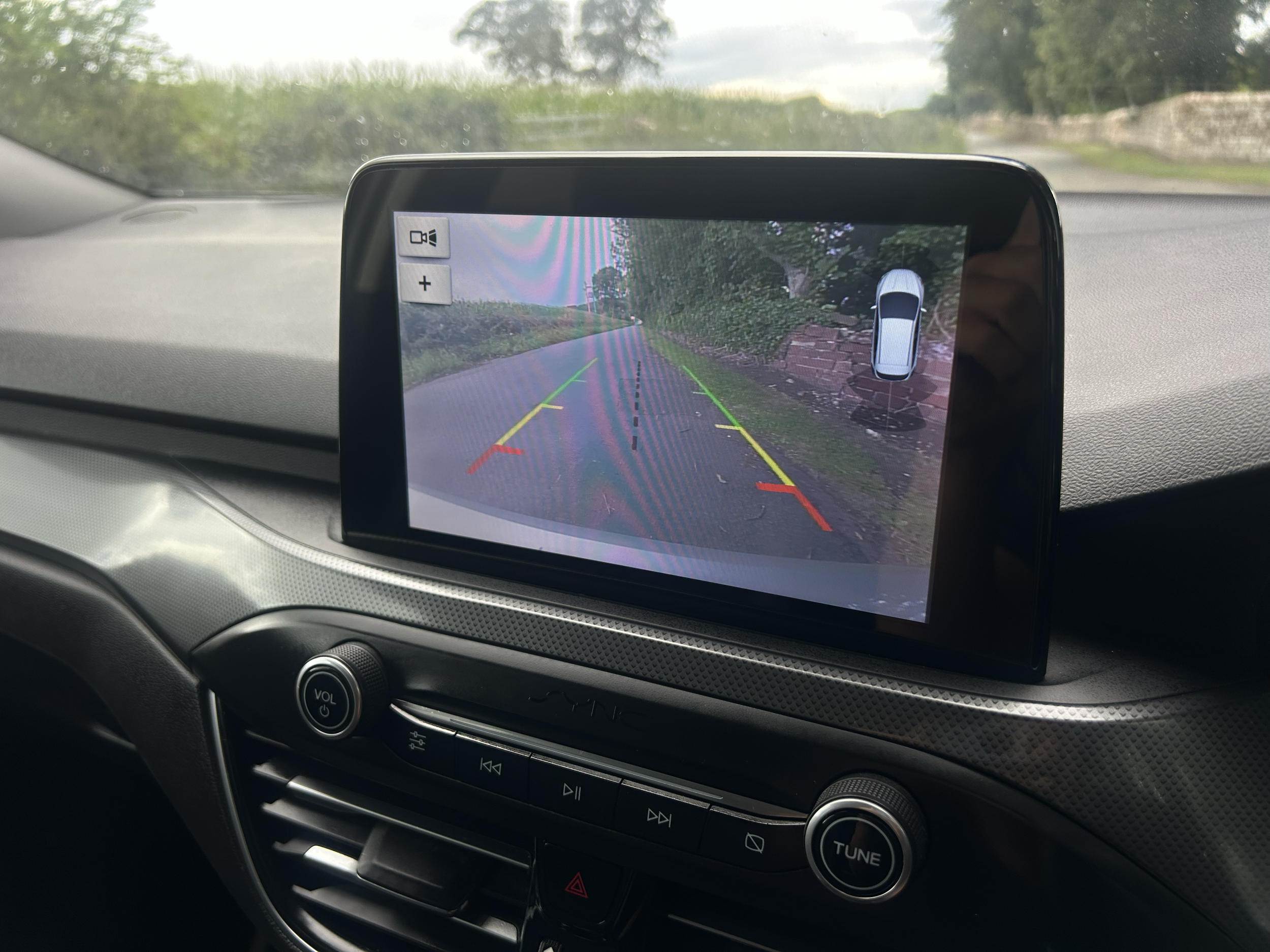 A car's dashboard with a touchscreen displaying a rearview camera feed showing a narrow rural road with greenery and trees, along with parking assist lines.