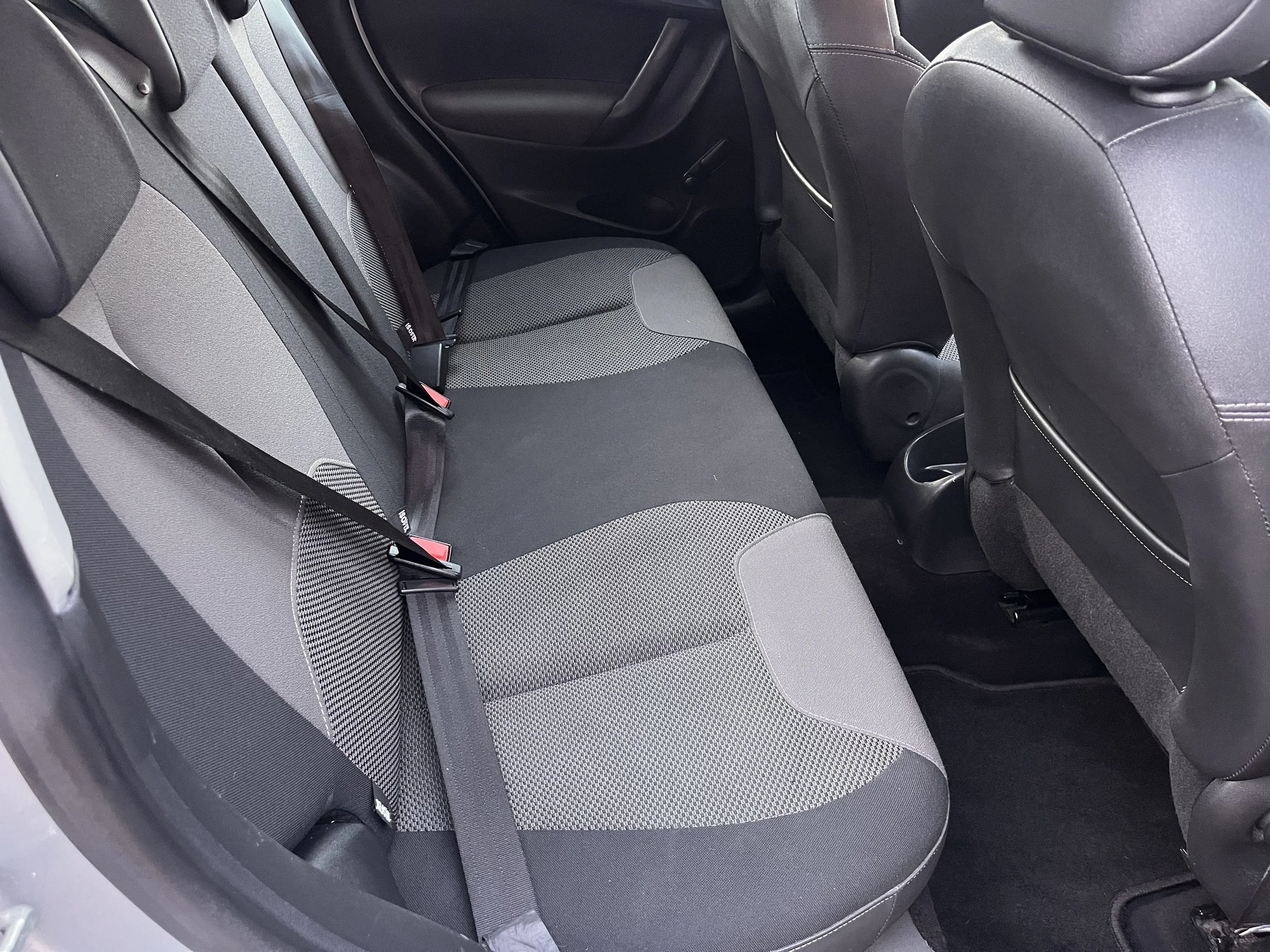 The interior of a car showing the backseat area with three seatbelts and black, gray, and patterned fabric upholstery.