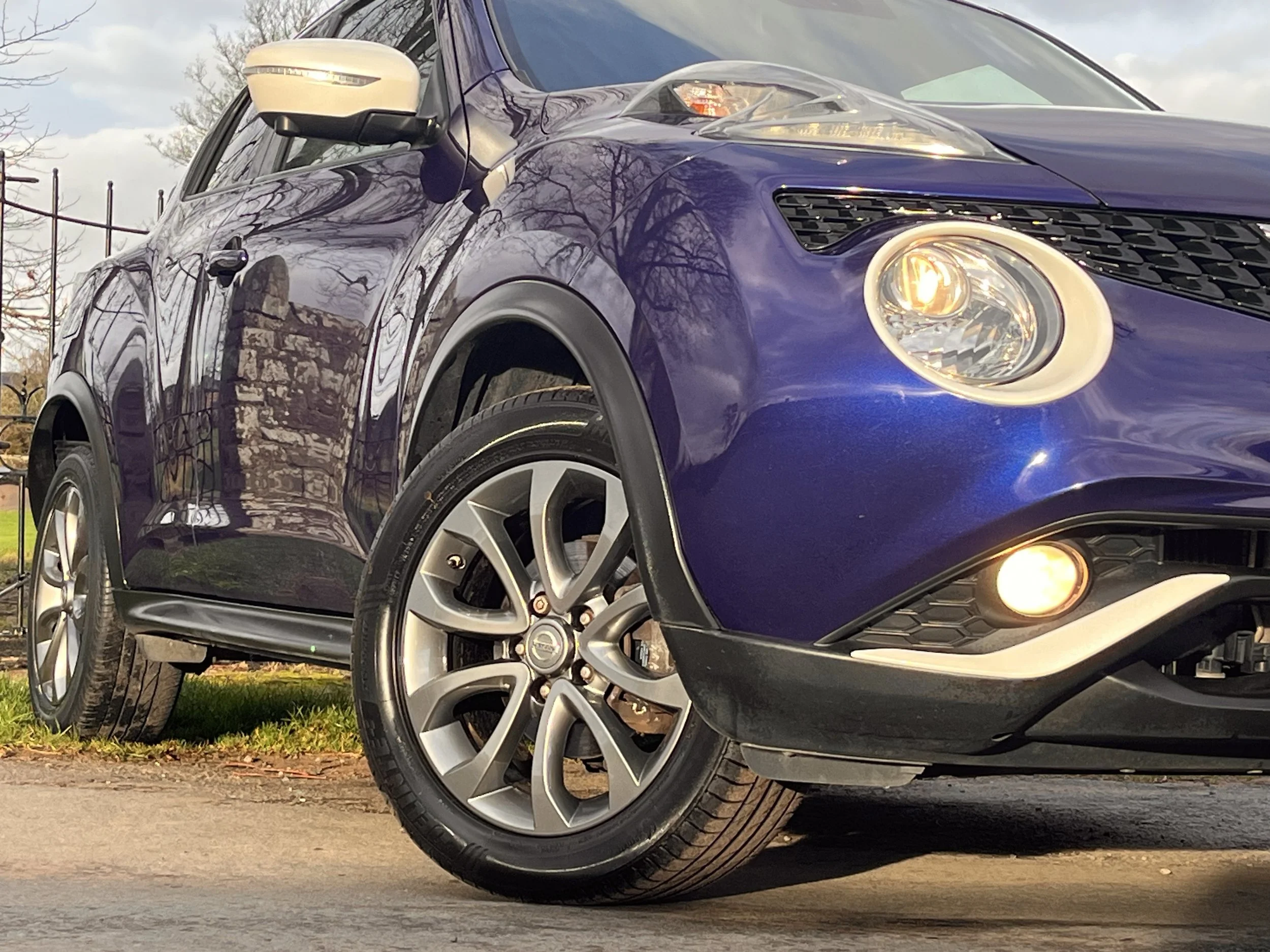 Close-up of a purple compact SUV showing front wheel, headlights, and side mirror, with a reflection of trees and sky on the car's body.