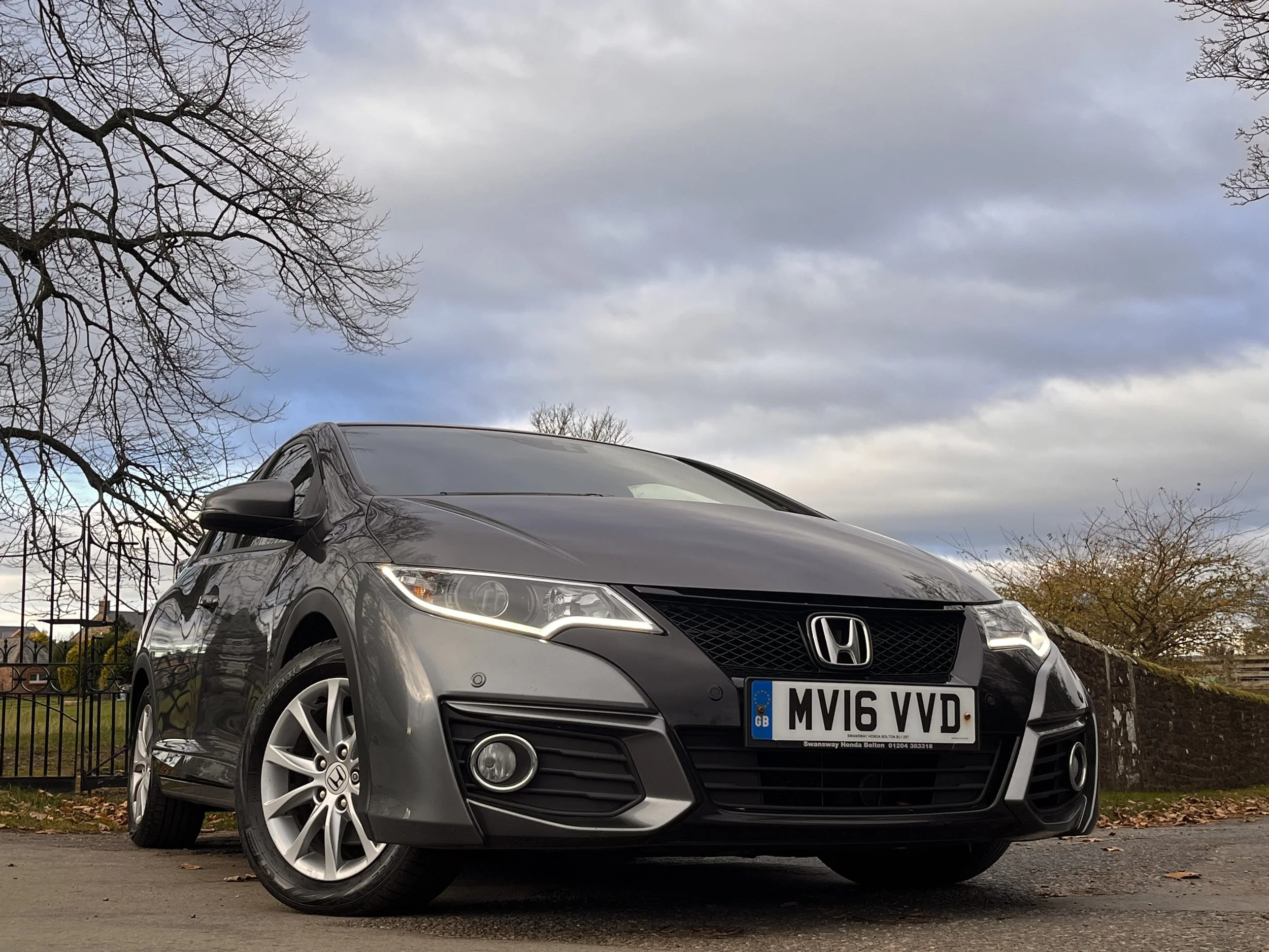 A black Honda Civic parked outdoors on a paved surface with a cloudy sky, leafless trees, and a brick wall in the background.