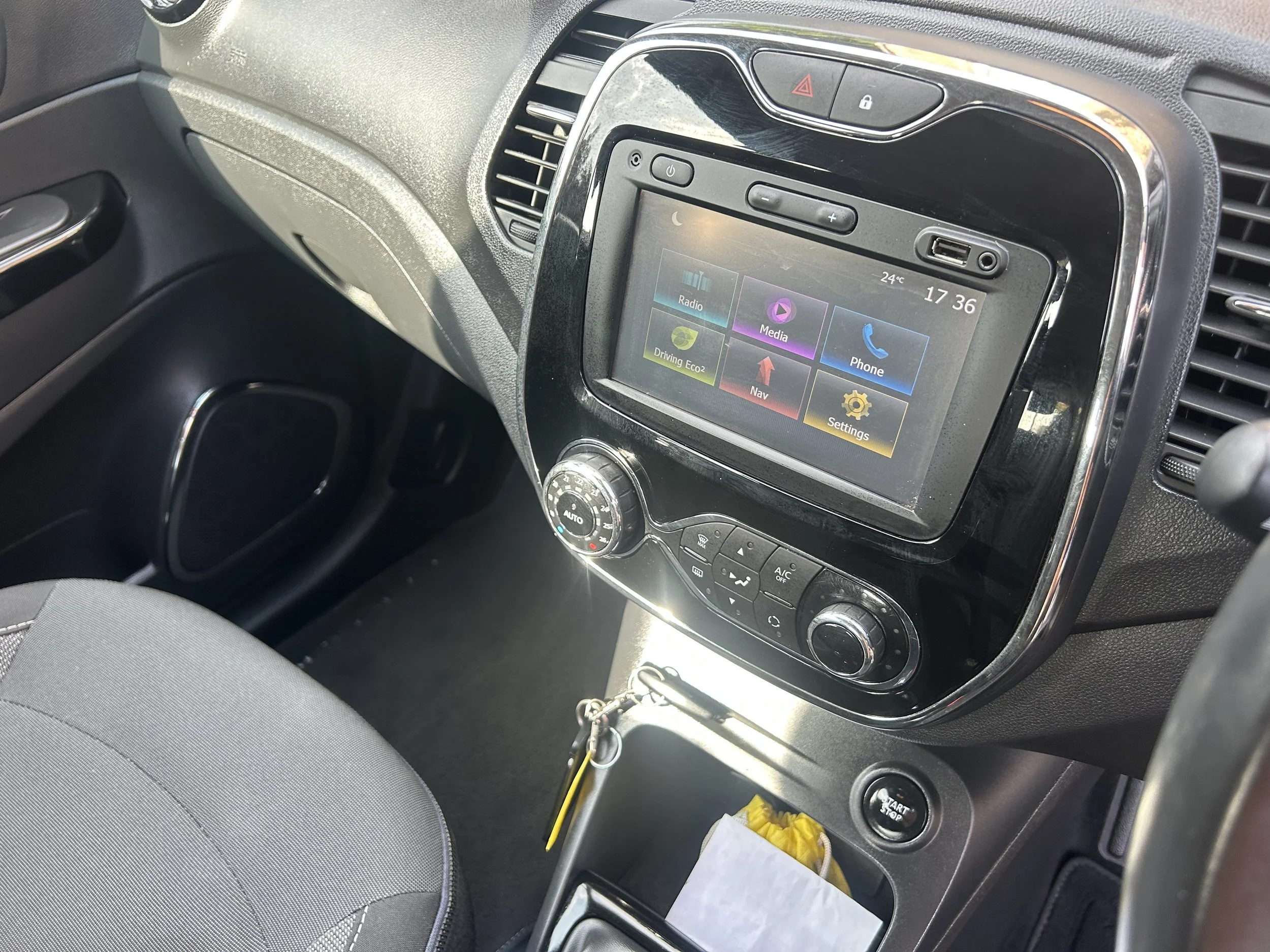 The photo shows the interior dashboard of a vehicle, including the central touchscreen display with options for radio, media, phone, driving eco, navigation, and settings. The dashboard has various control buttons, dials, a start/stop button, and a k