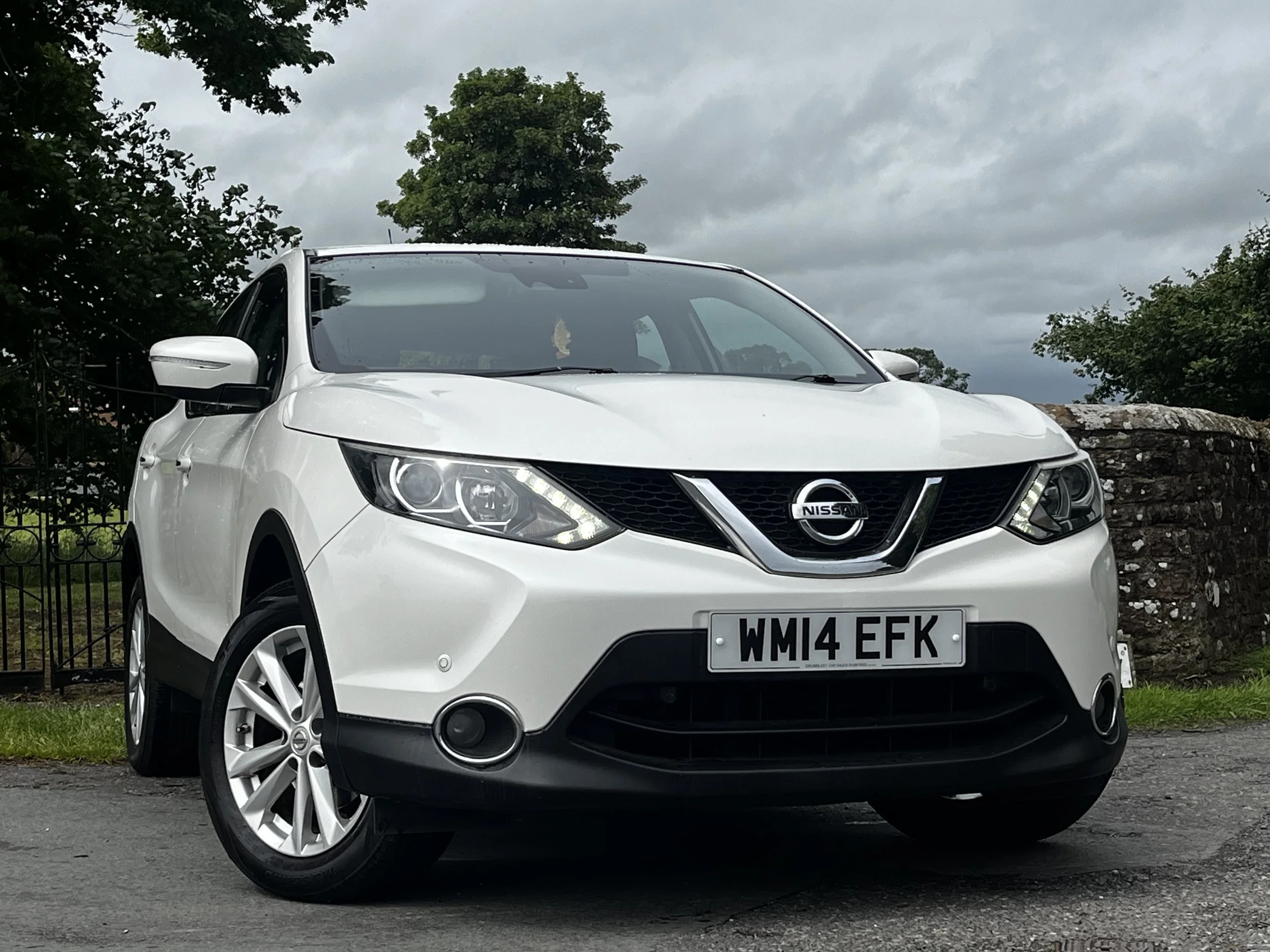 A white Nissan car with a UK license plate WM14 EFK parked outdoors on a paved surface. The background features green trees, a stone wall, and cloudy sky.