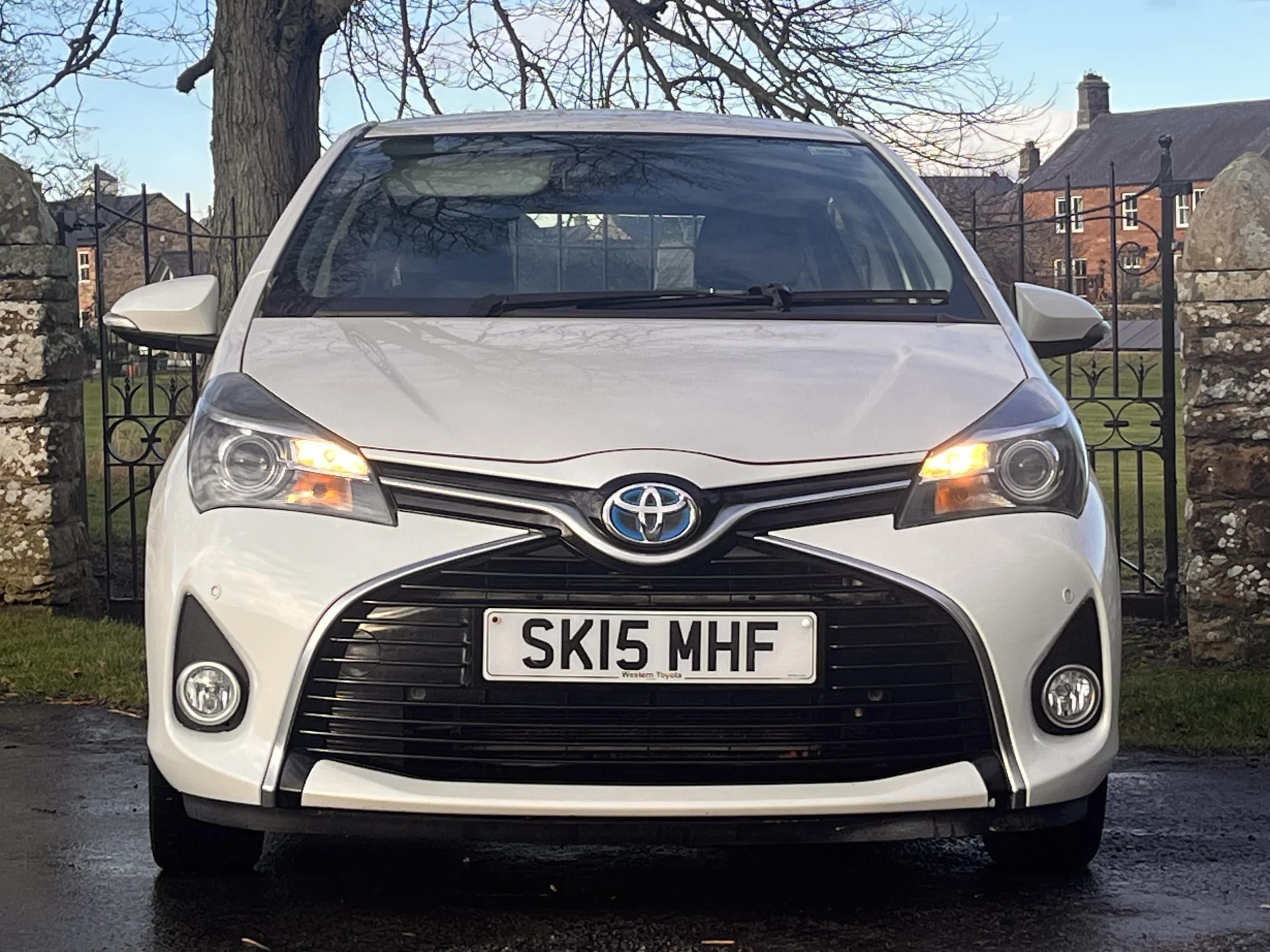 Front view of a white Toyota car with a UK license plate 'SK15 MHF', parked outdoors with trees and houses in the background.