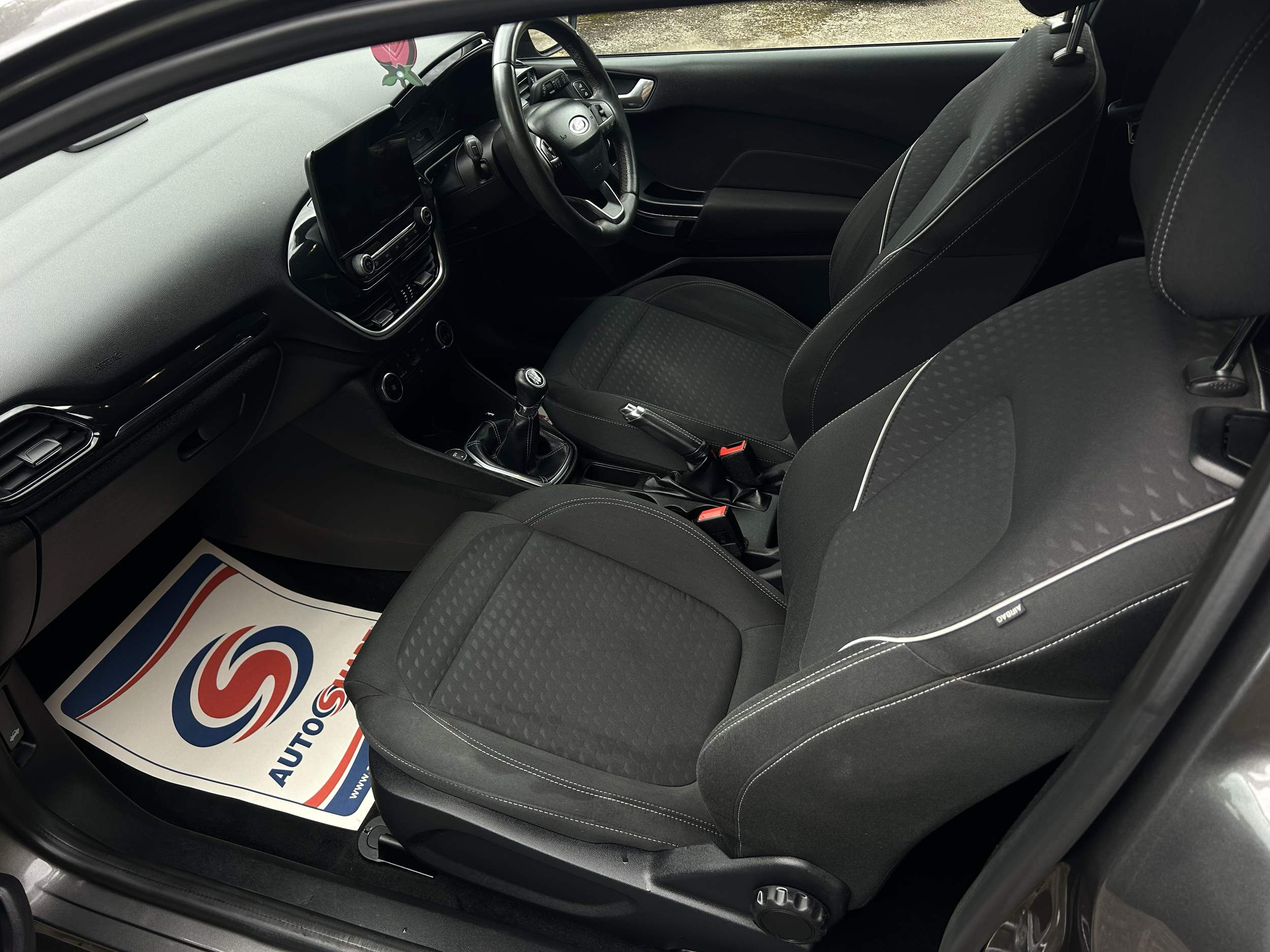 Interior view of a car showing the front passenger seat, dashboard, steering wheel, and a floor mat with a logo.