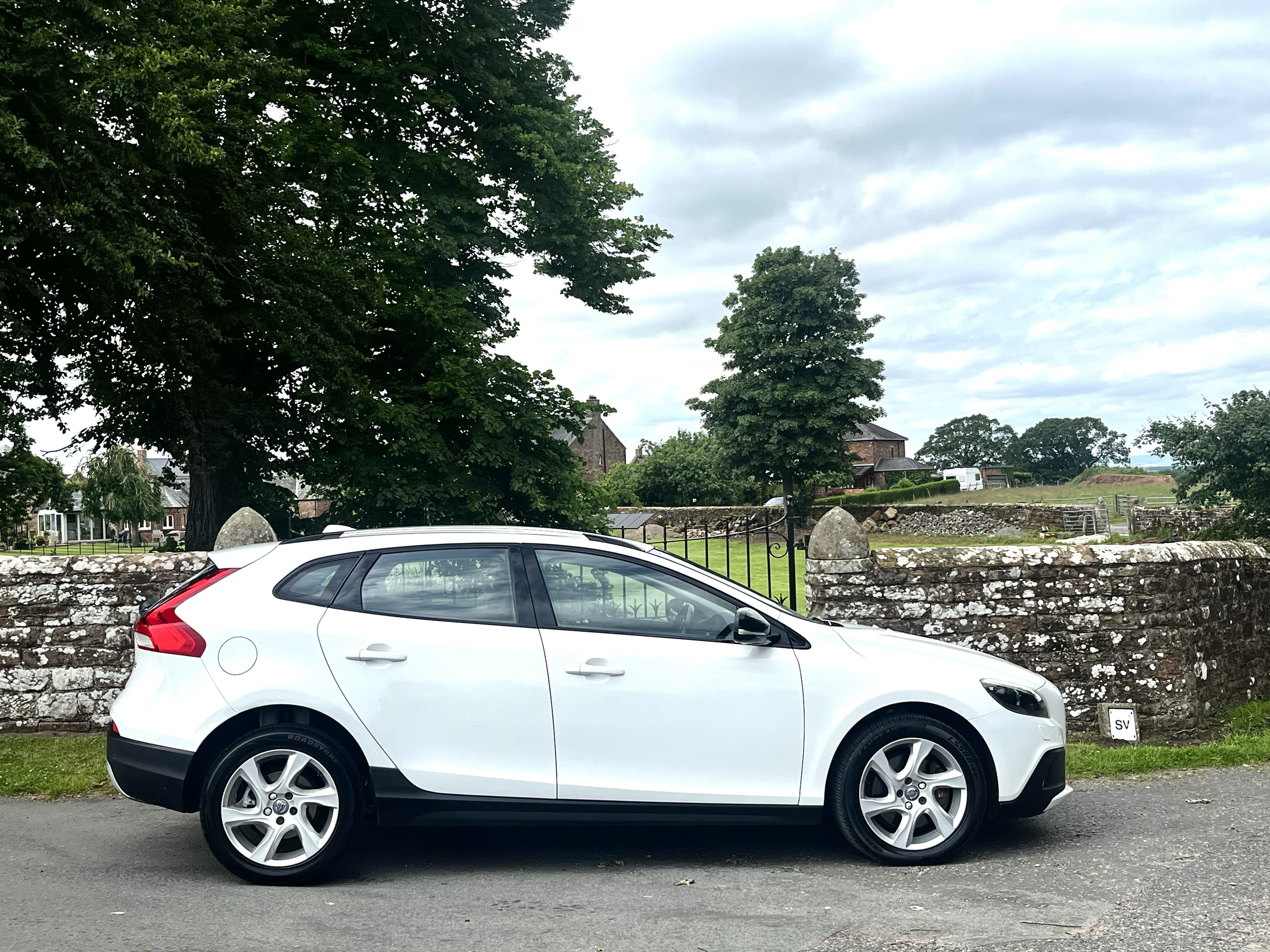 White compact crossover vehicle parked on a street in front of a stone wall and green grass, with trees, houses, and cloudy sky in the background.