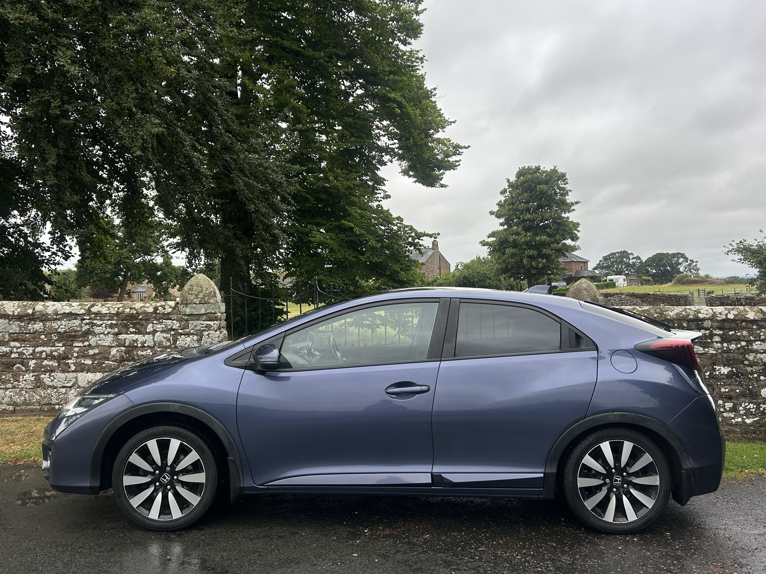 A silver sedan car parked on the street in front of a stone wall, with large trees and old buildings in the background on a cloudy day.