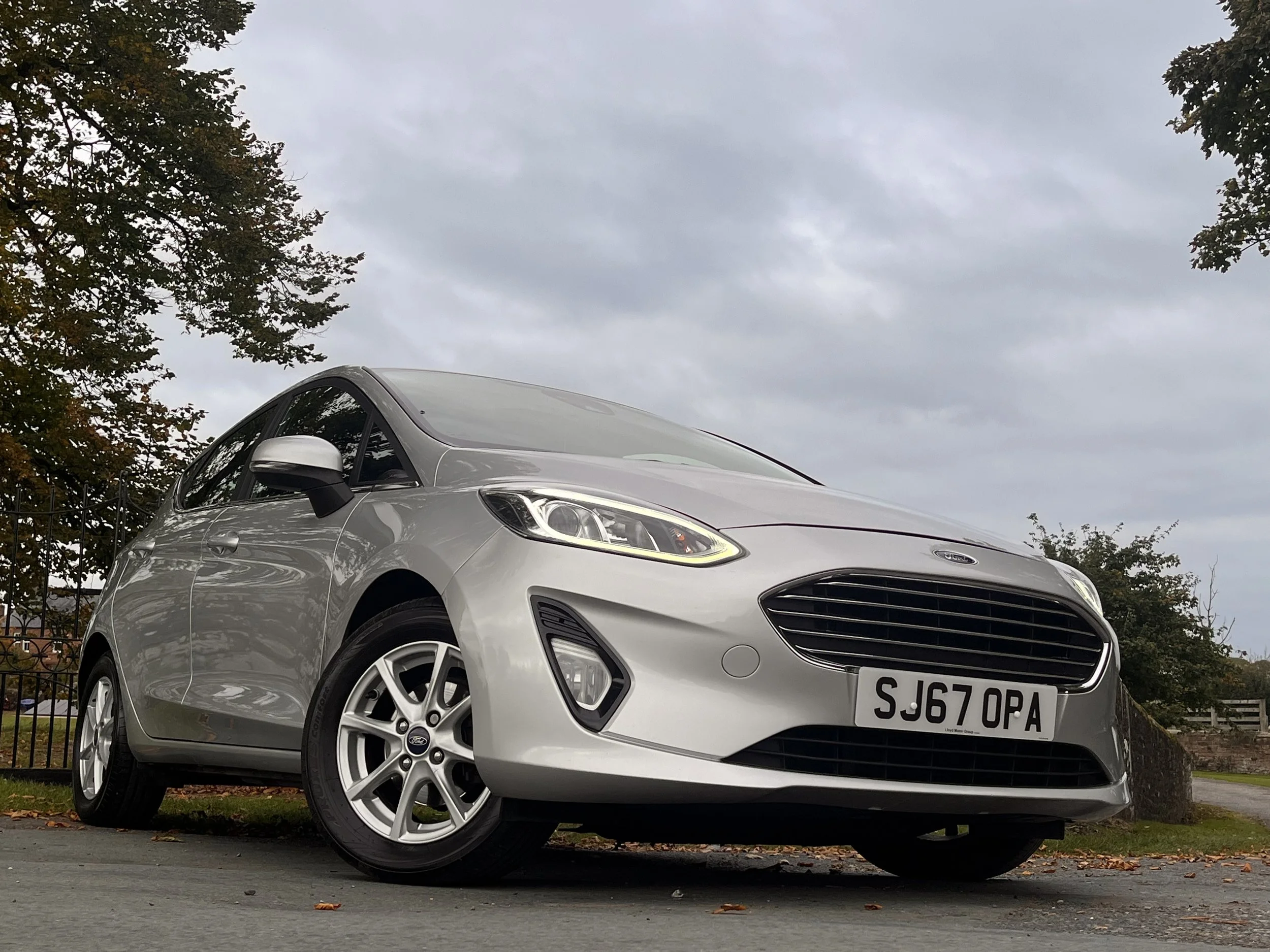 Silver Ford Fiesta parked on the street with trees and cloudy sky in the background.