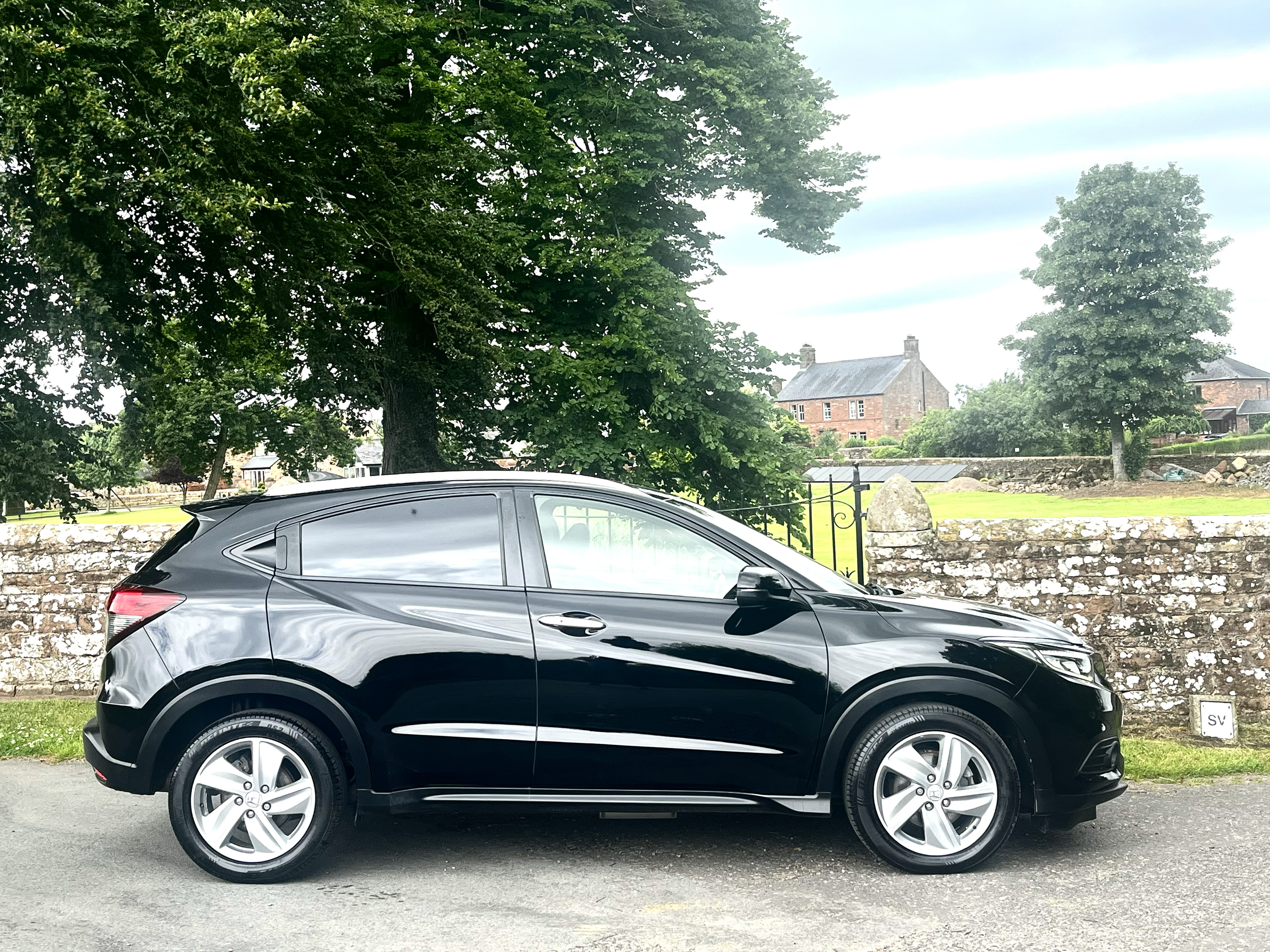 A black SUV parked on a street with a brick wall and a grassy area in the background, trees and houses visible in the distance, under a partly cloudy sky.