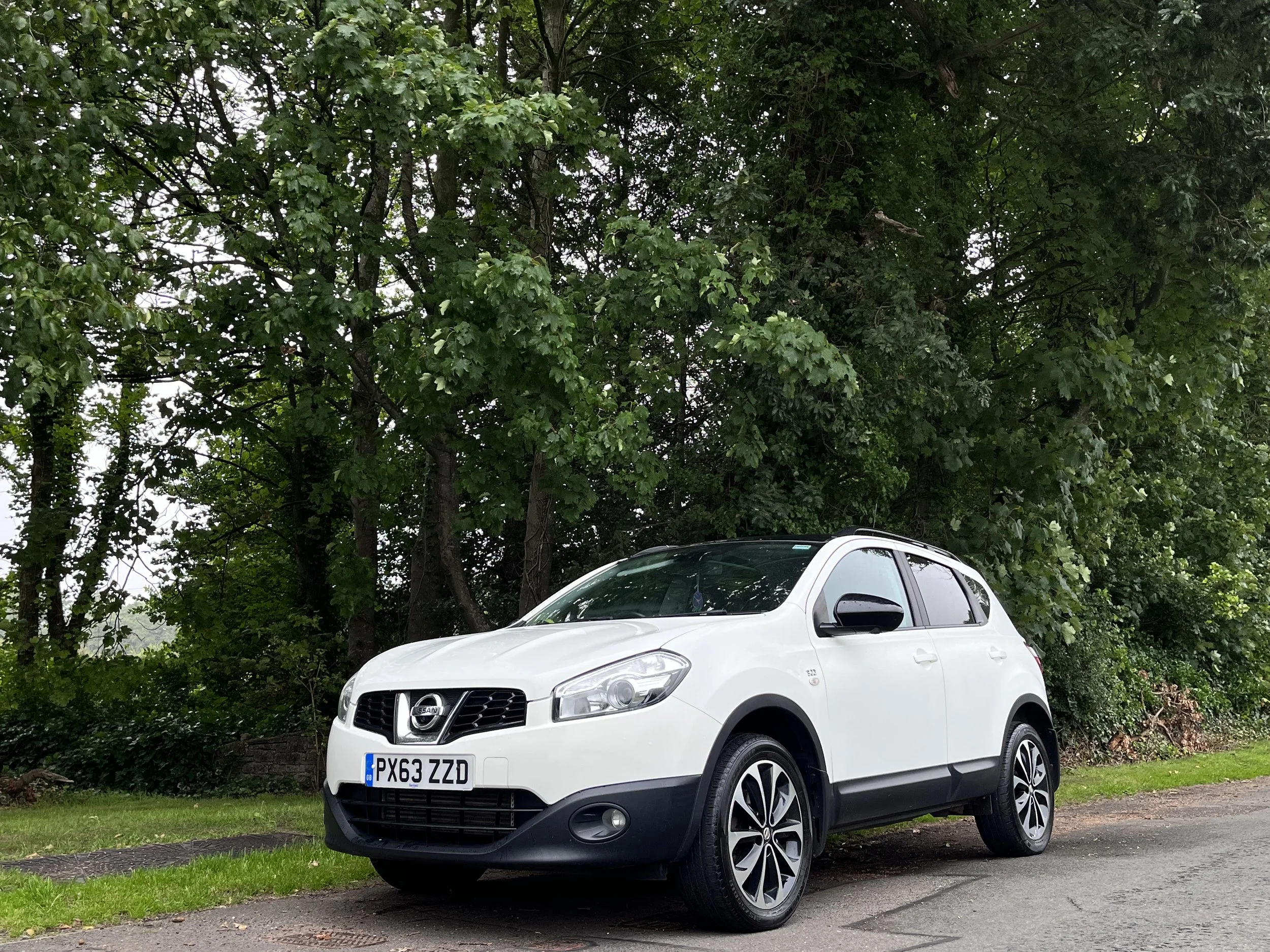 White Nissan SUV parked on the side of a road with green trees in the background.