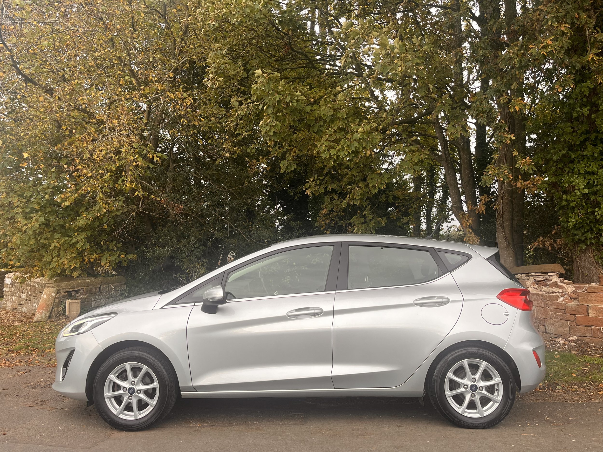 Silver hatchback car parked on the side of the street with large green trees and a brick wall in the background.
