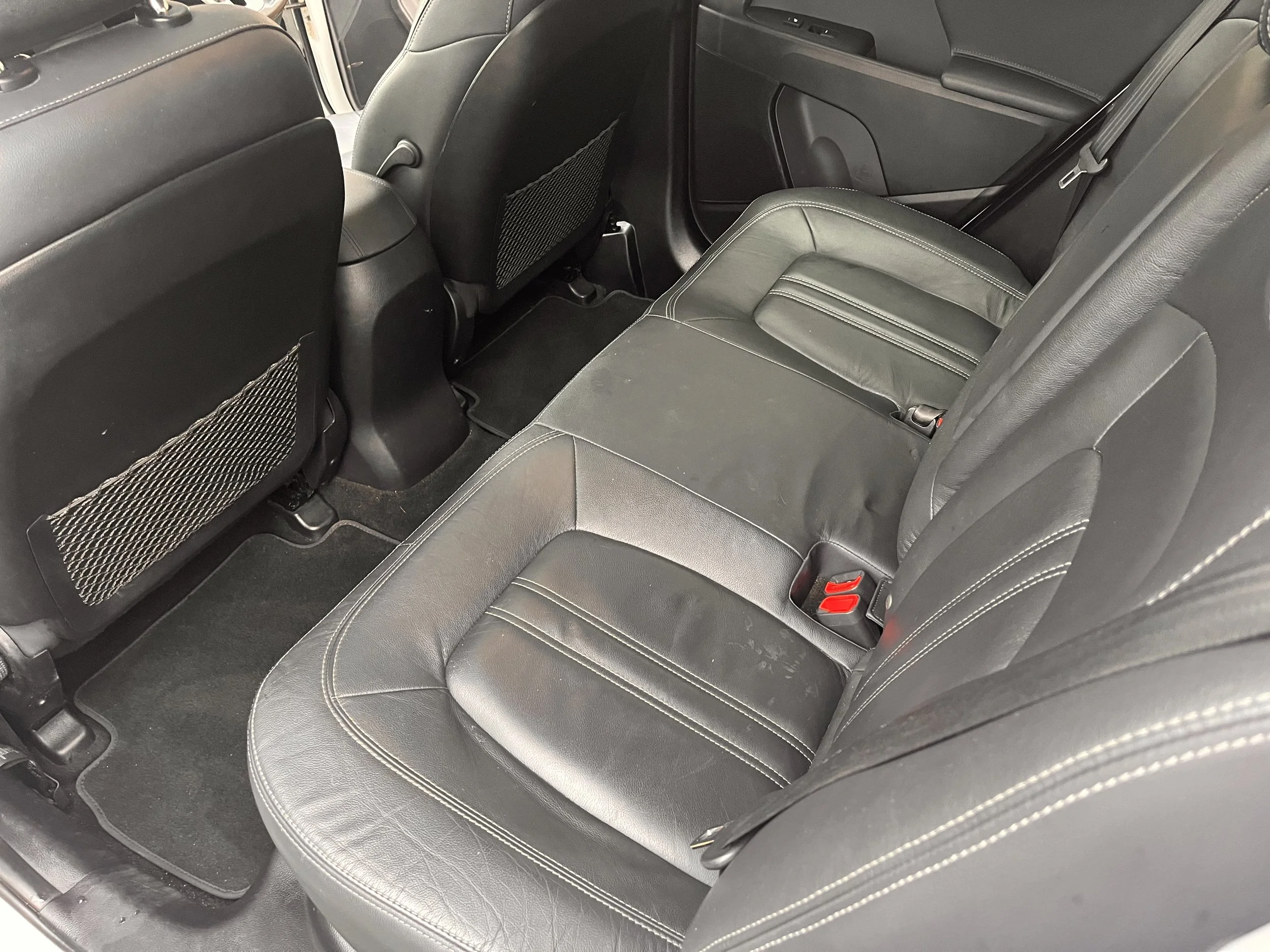 The interior of a car showing the backseat area with black leather seats and floor mats. The back of the front seats has mesh storage pockets.