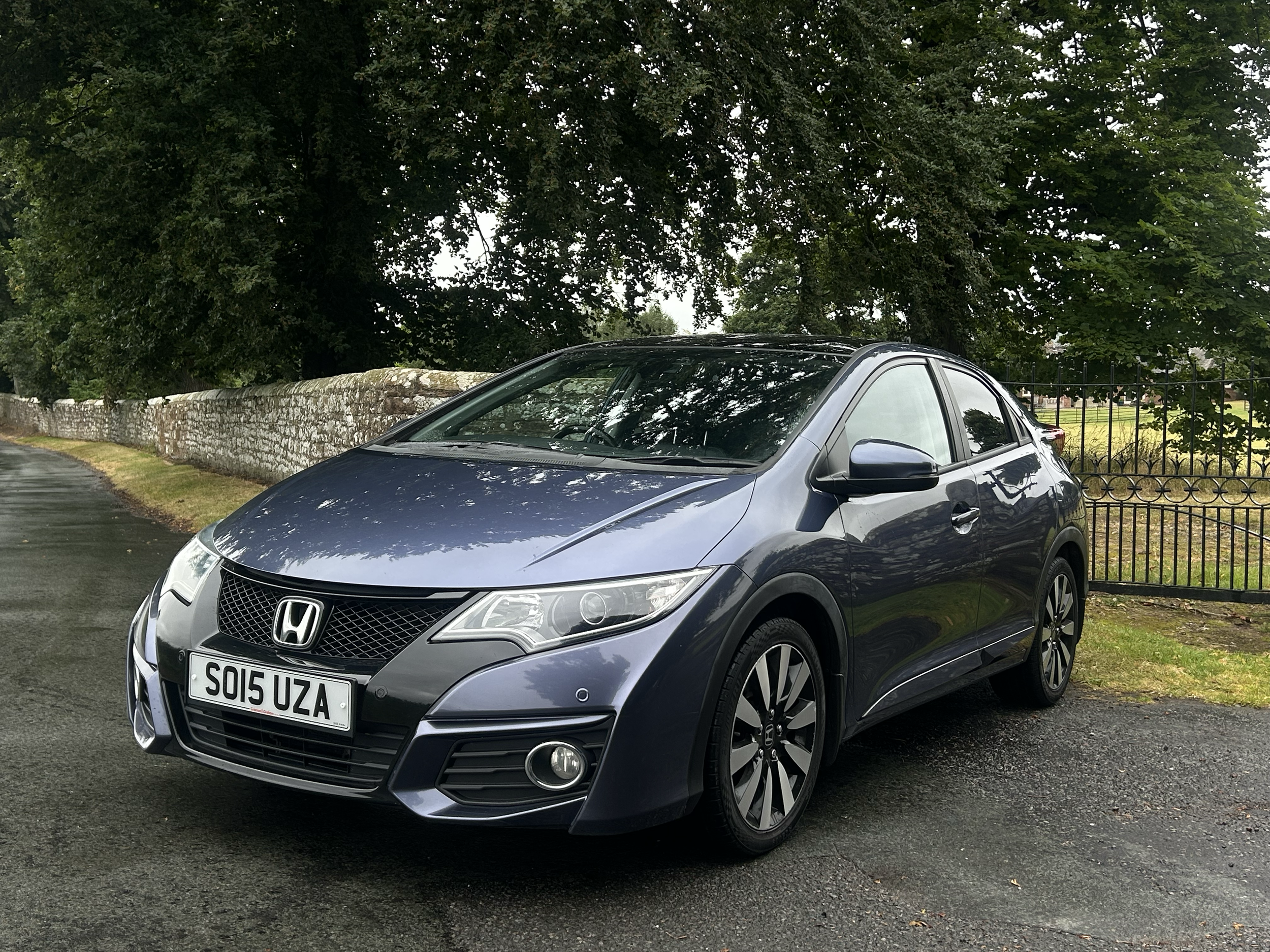 A metallic gray Honda car parked on a street with a stone wall and large leafy trees in the background.
