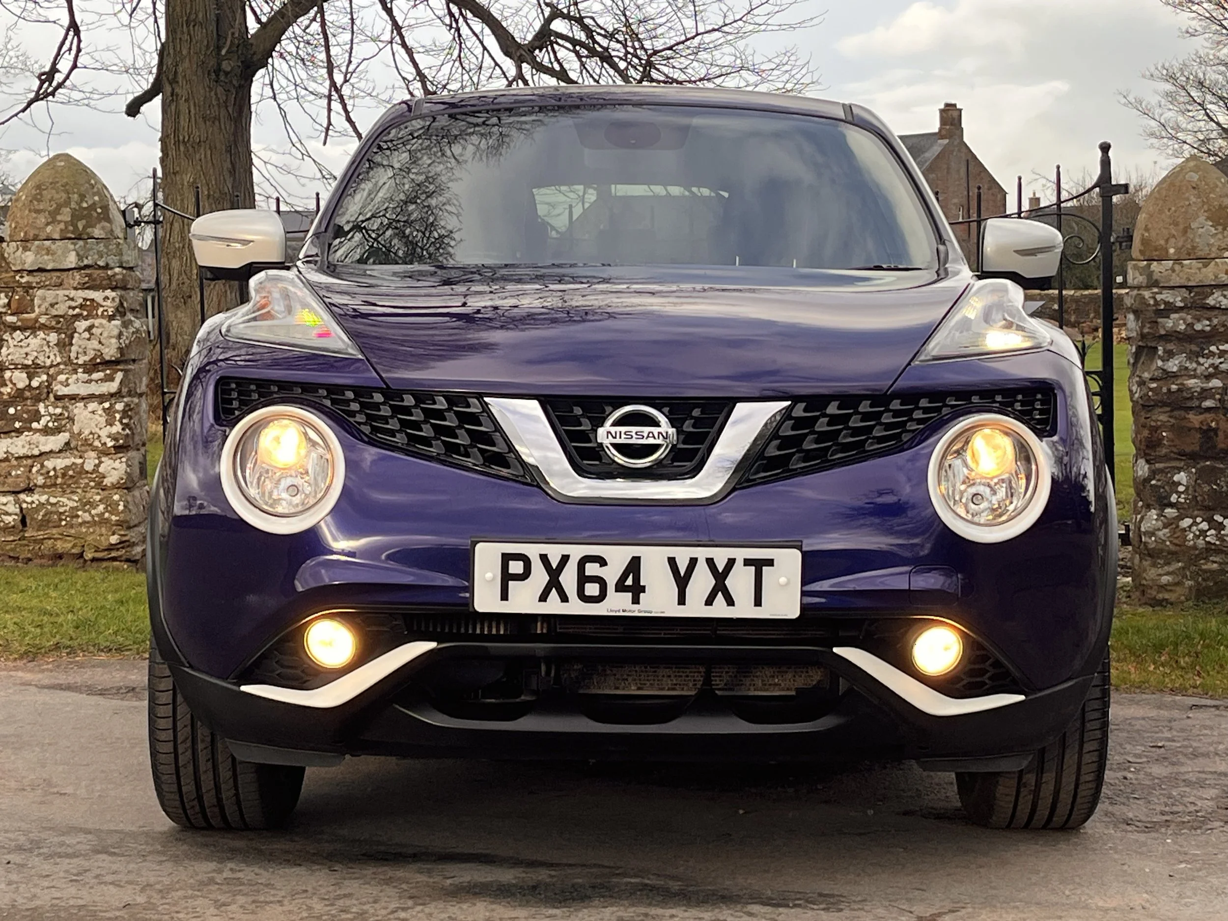 Front view of a purple Nissan Juke with headlights and fog lights on, parked in front of stone pillars and a rural landscape with trees and a house in the background.