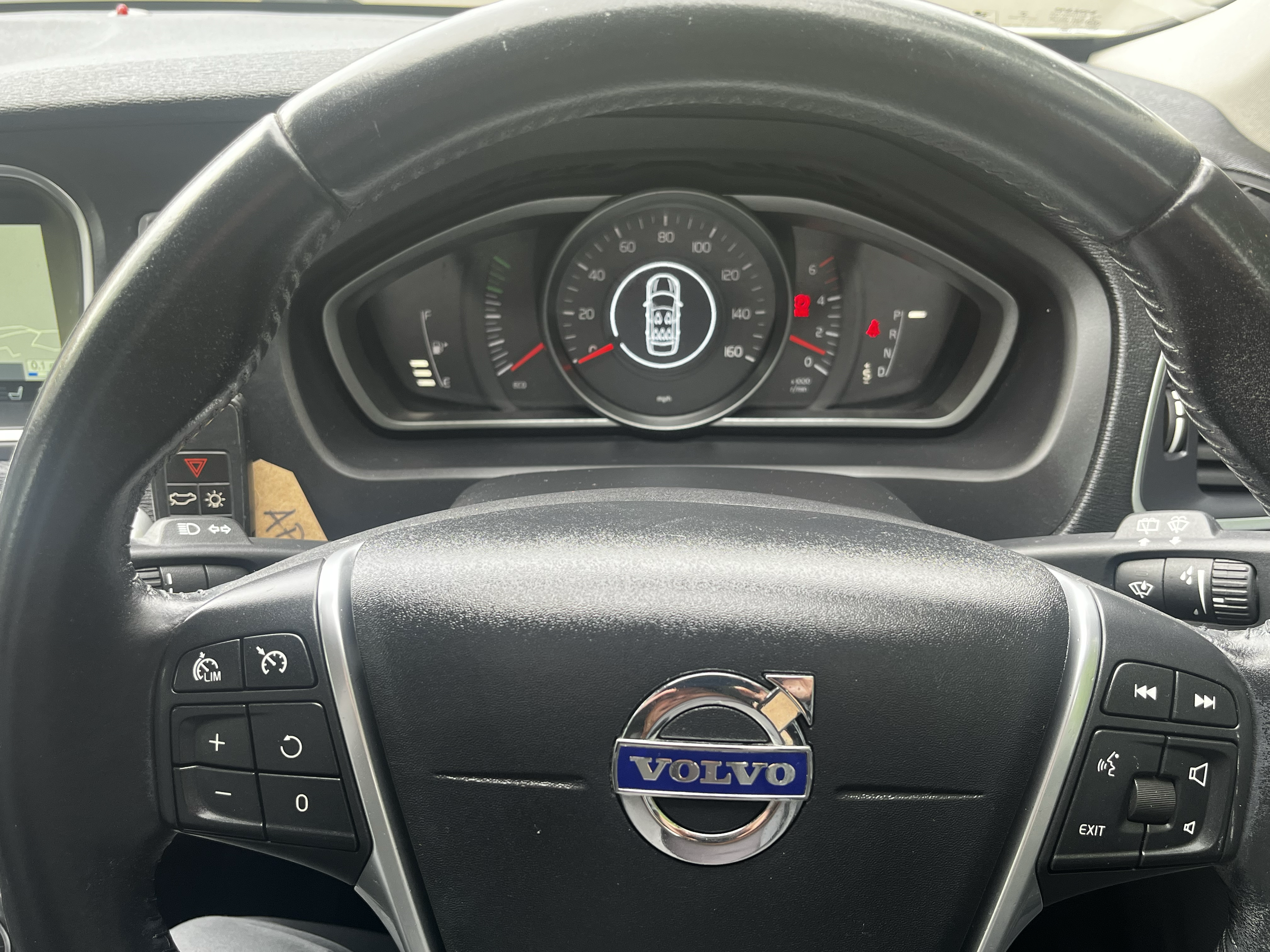 Interior view of a Volvo car dashboard with a steering wheel that displays the Volvo logo, speedometer, fuel gauge, and various control buttons.
