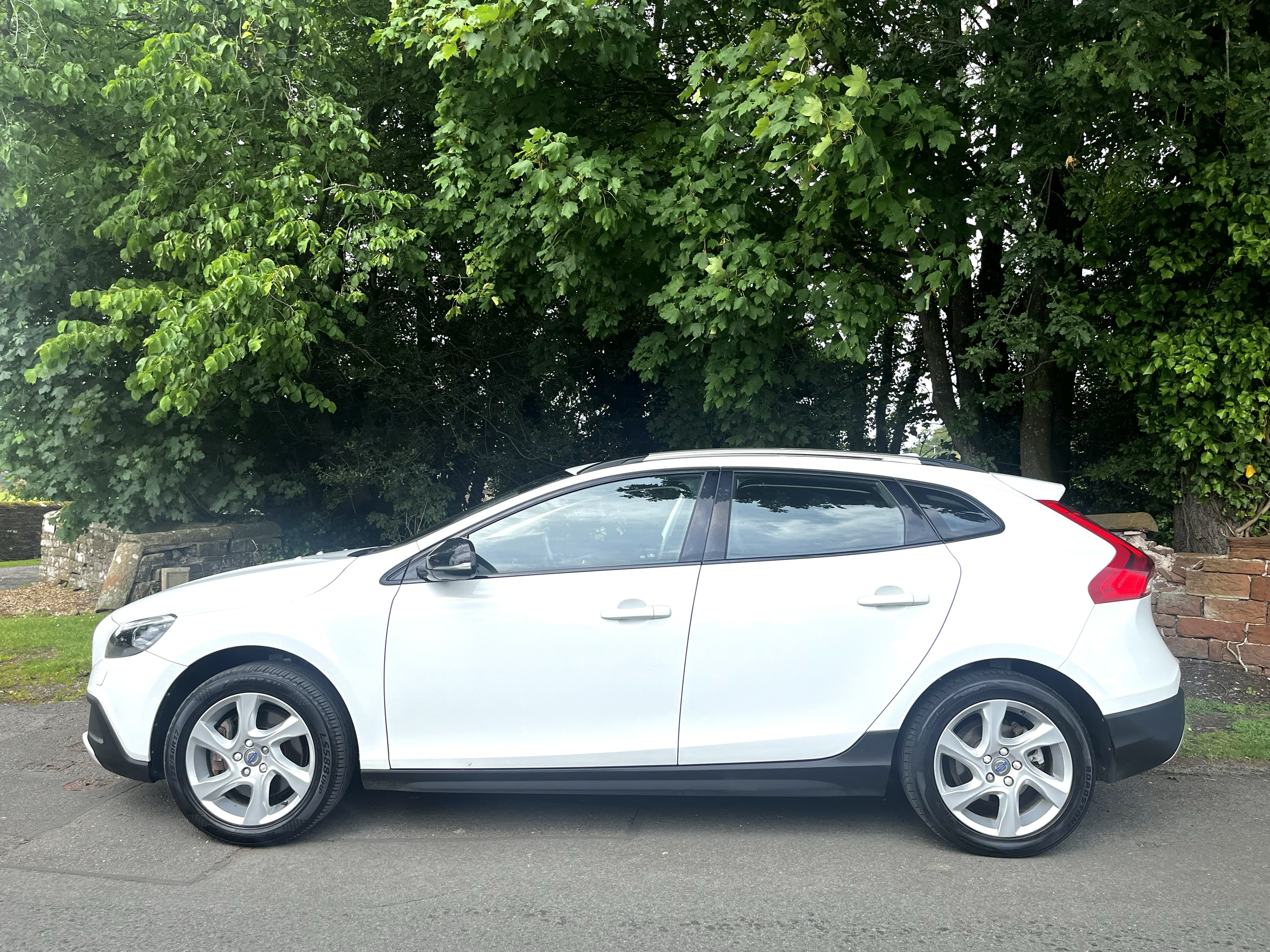 White modern hatchback car parked on the street in front of green leafy trees and under cloudy sky.