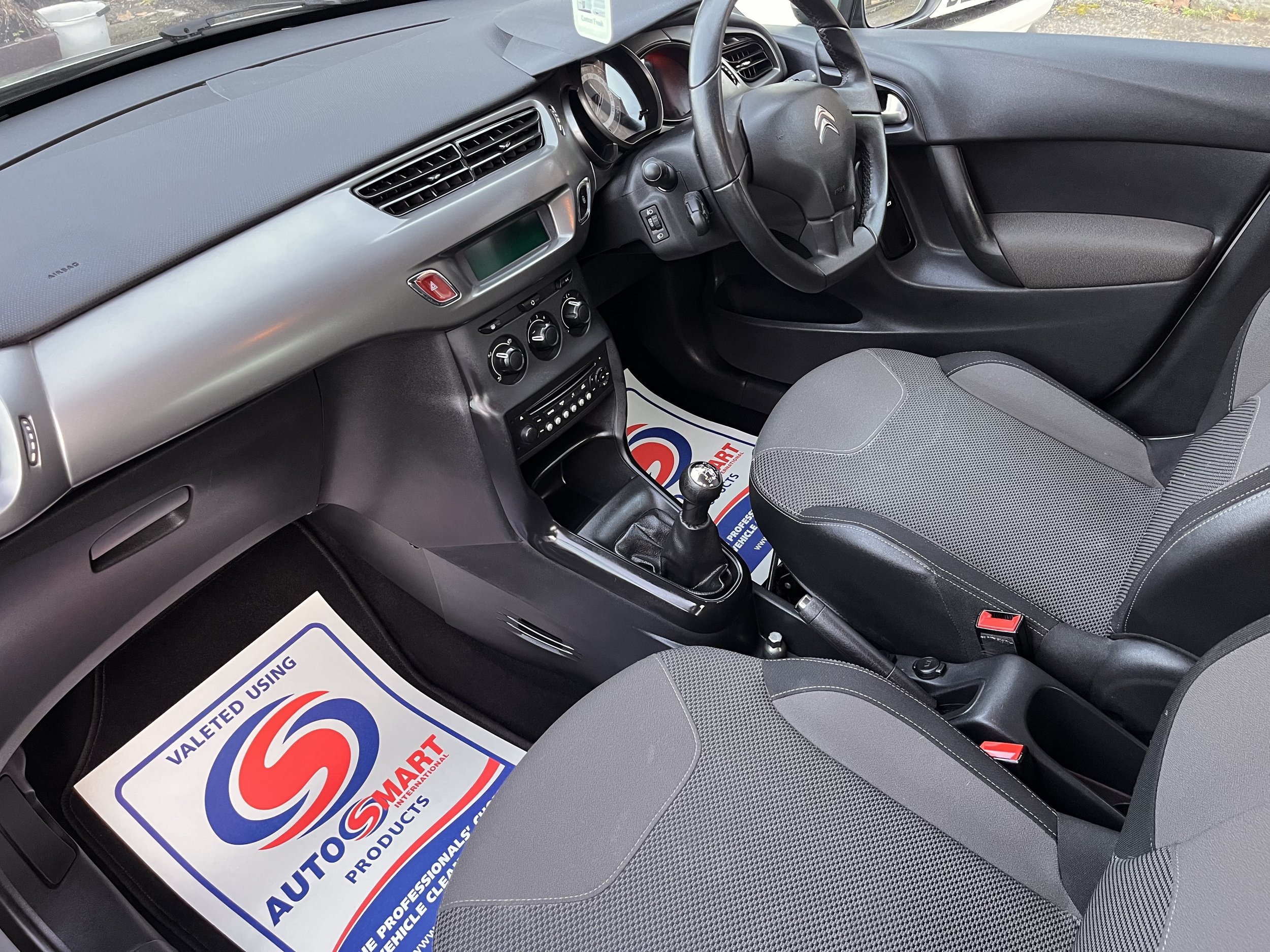 Interior of a car with a manual transmission, black and gray dashboard, two front seats with gray fabric upholstery, steering wheel, gear shift, and floor mats.