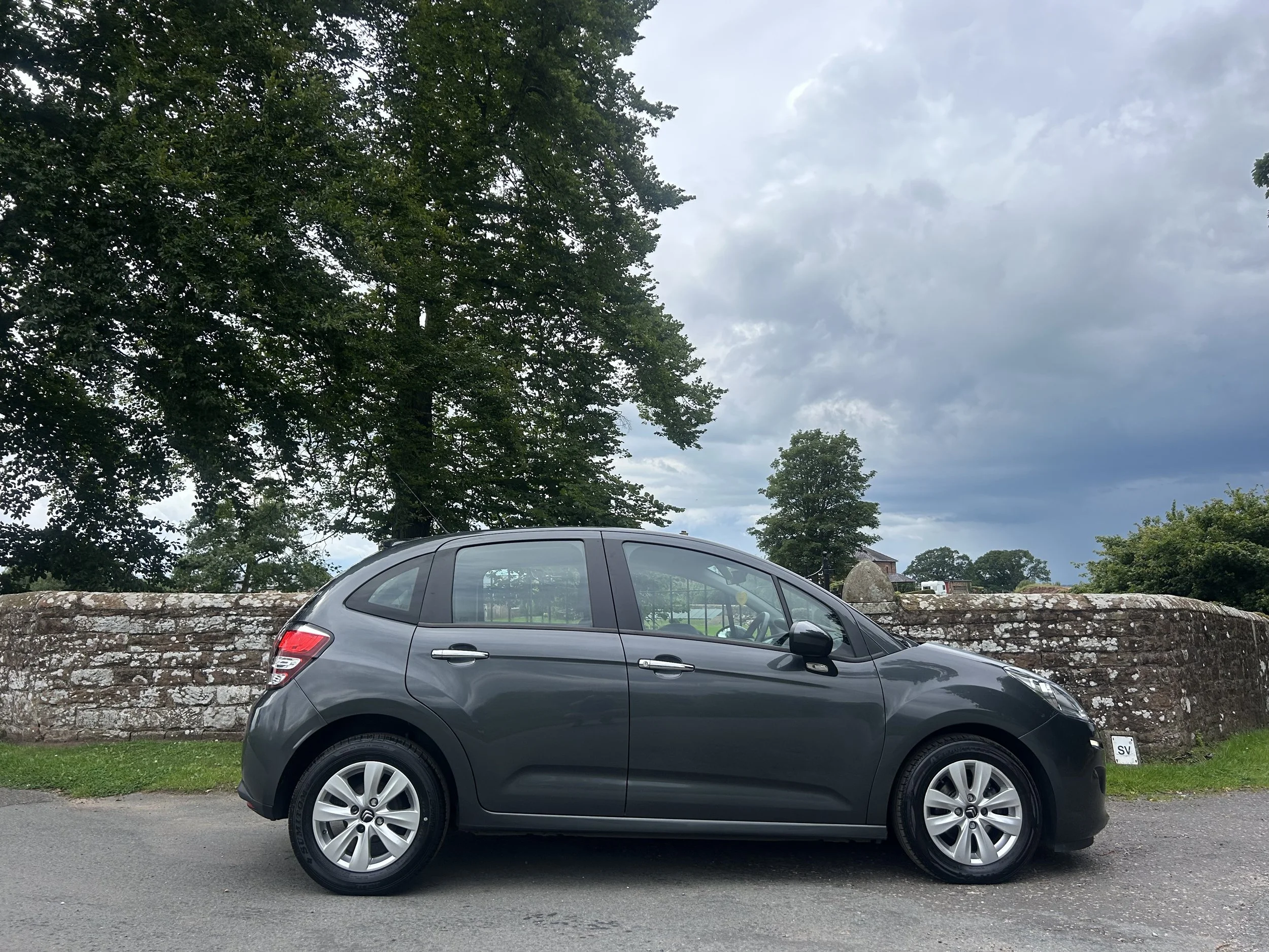 Gray hatchback car parked on road next to a stone wall with trees and cloudy sky in the background.