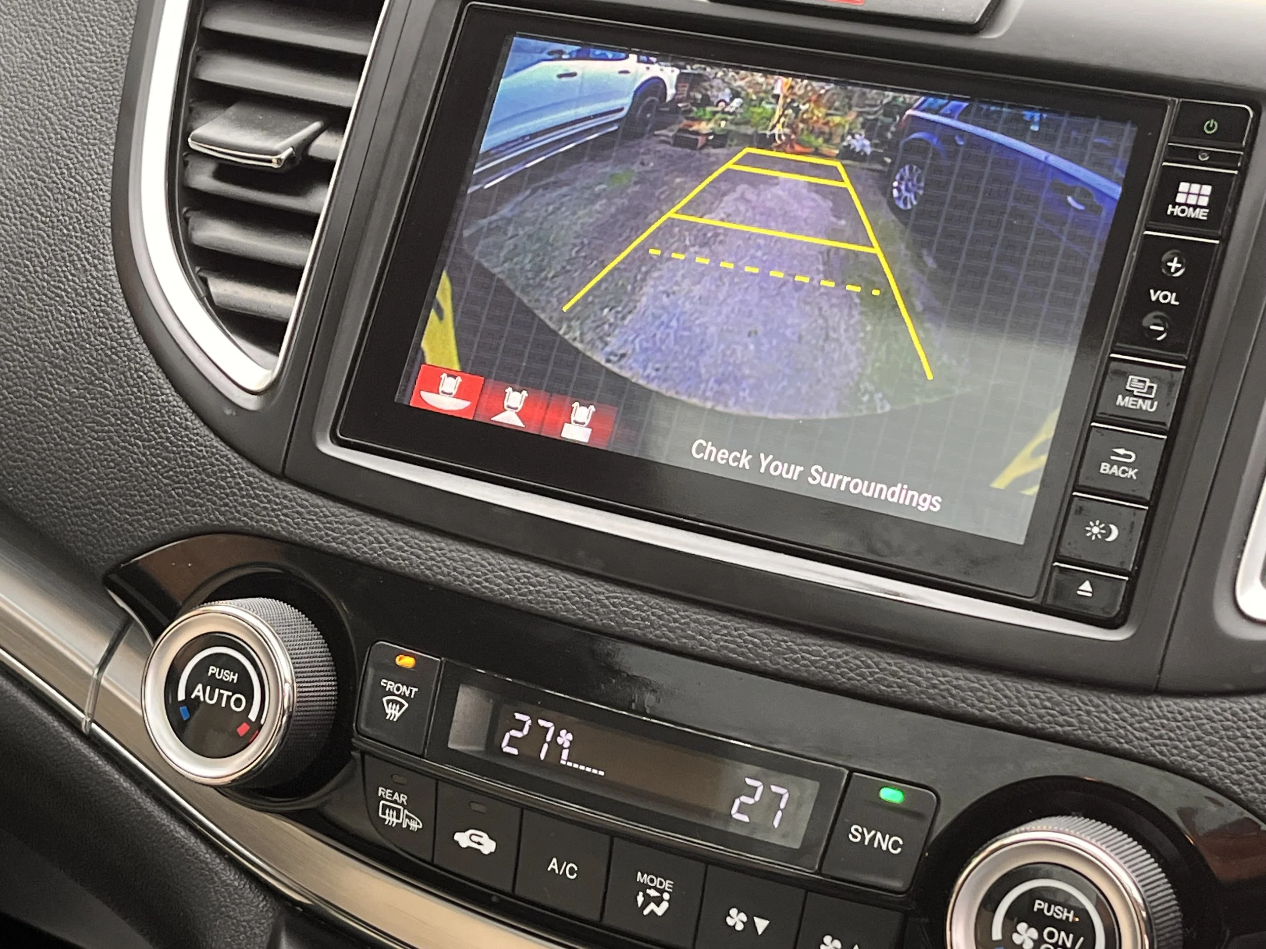 The image shows a car's dashboard with a rearview camera display. The display indicates the view behind the vehicle, with yellow guiding lines and a message to check surroundings. The climate control system below shows a temperature of 27 degrees Cel