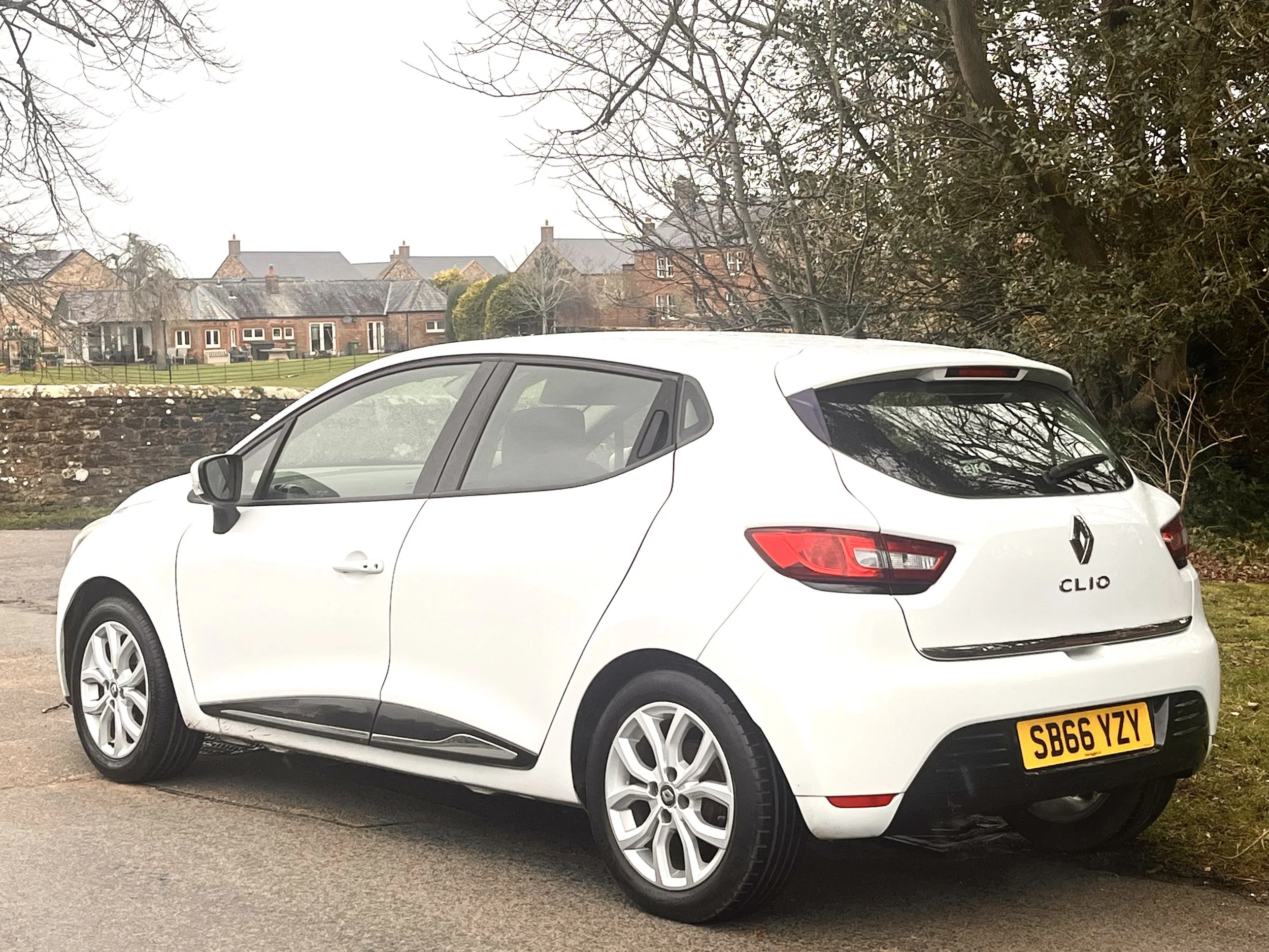 White Renault Clio parked on the side of the road with houses and trees in the background.