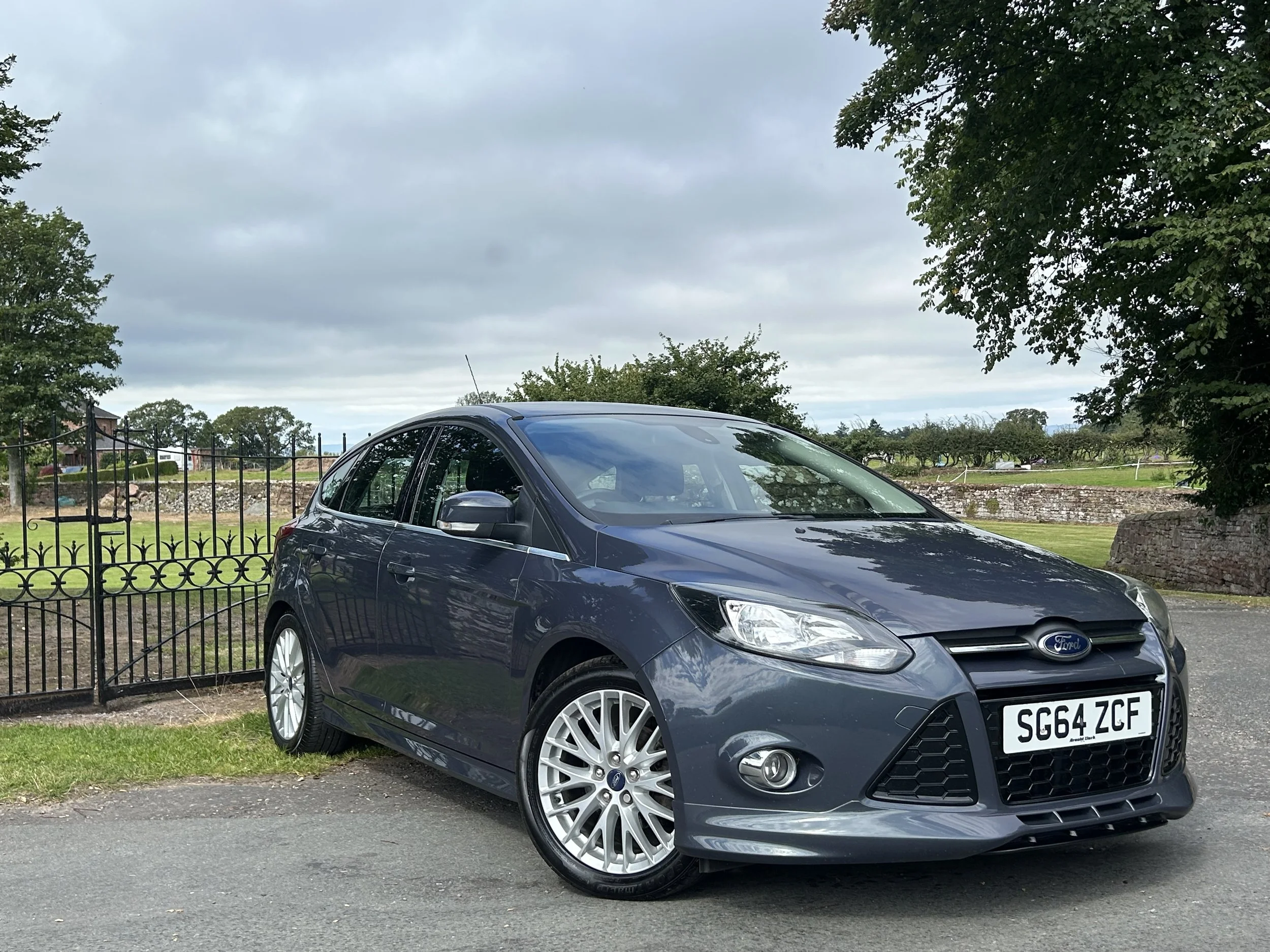 Gray Ford Focus parked on a street, with trees and a stone wall and fence in the background under cloudy skies.