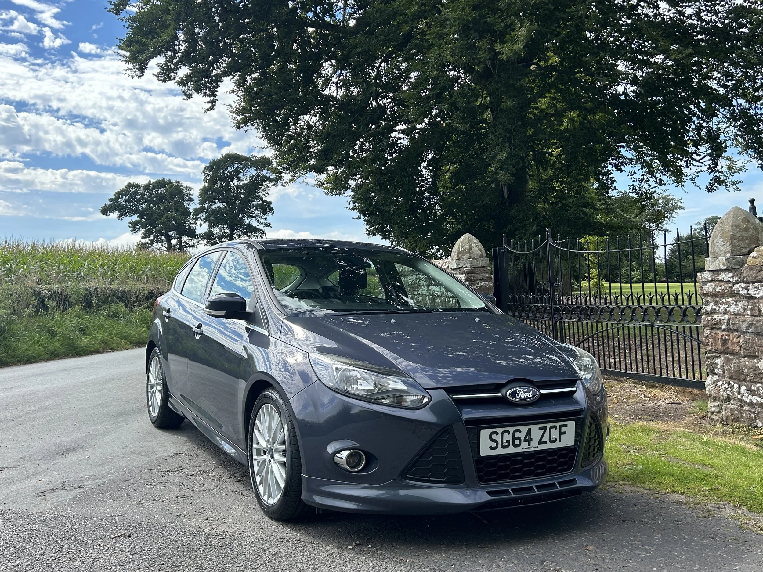 A dark gray Ford hatchback car parked on a rural road with large trees and a black iron gate in the background, under partly cloudy skies.