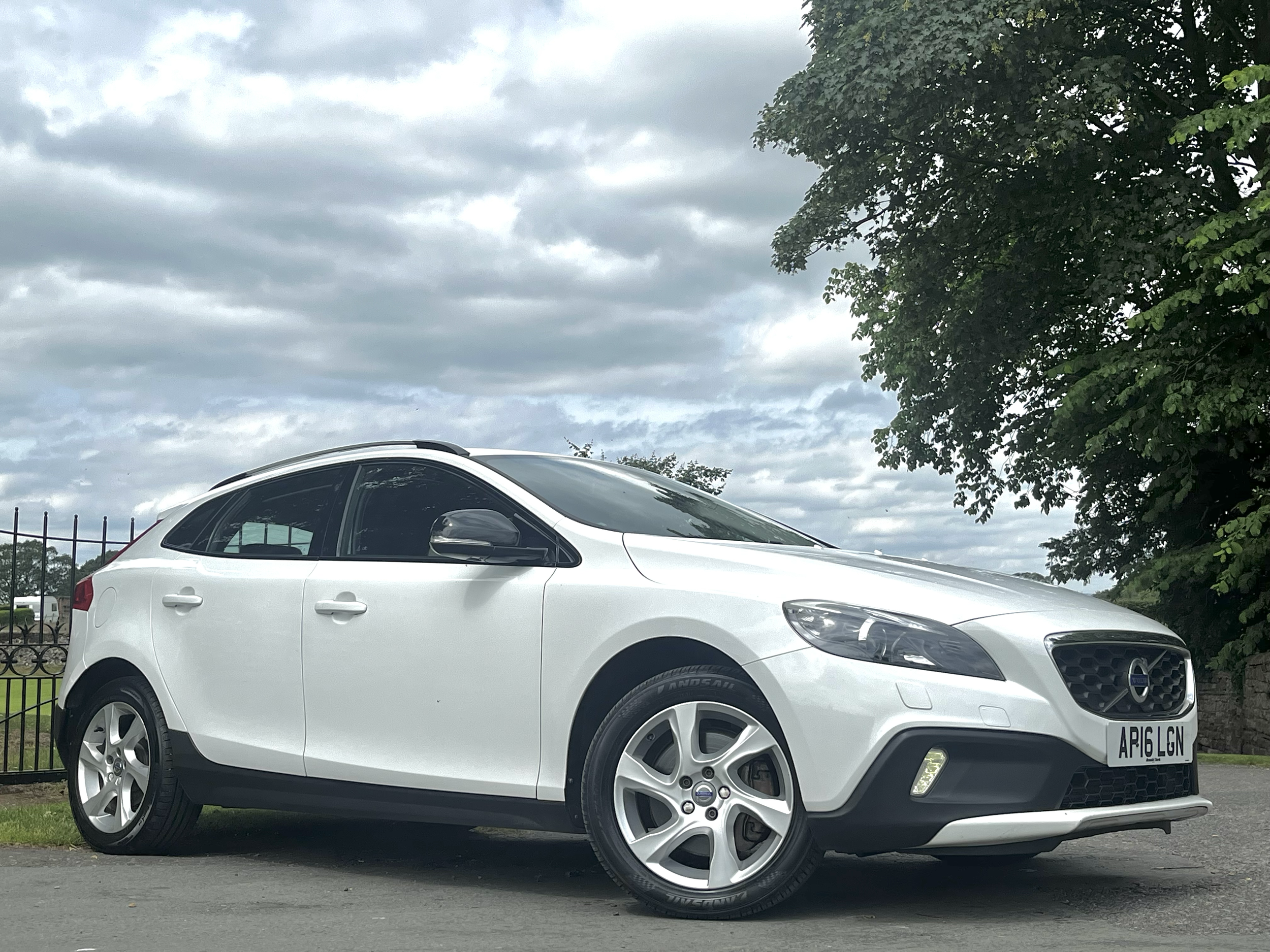 A white hatchback car parked on the side of the road with a black metal fence in the background, green trees, and an overcast sky filled with gray clouds.