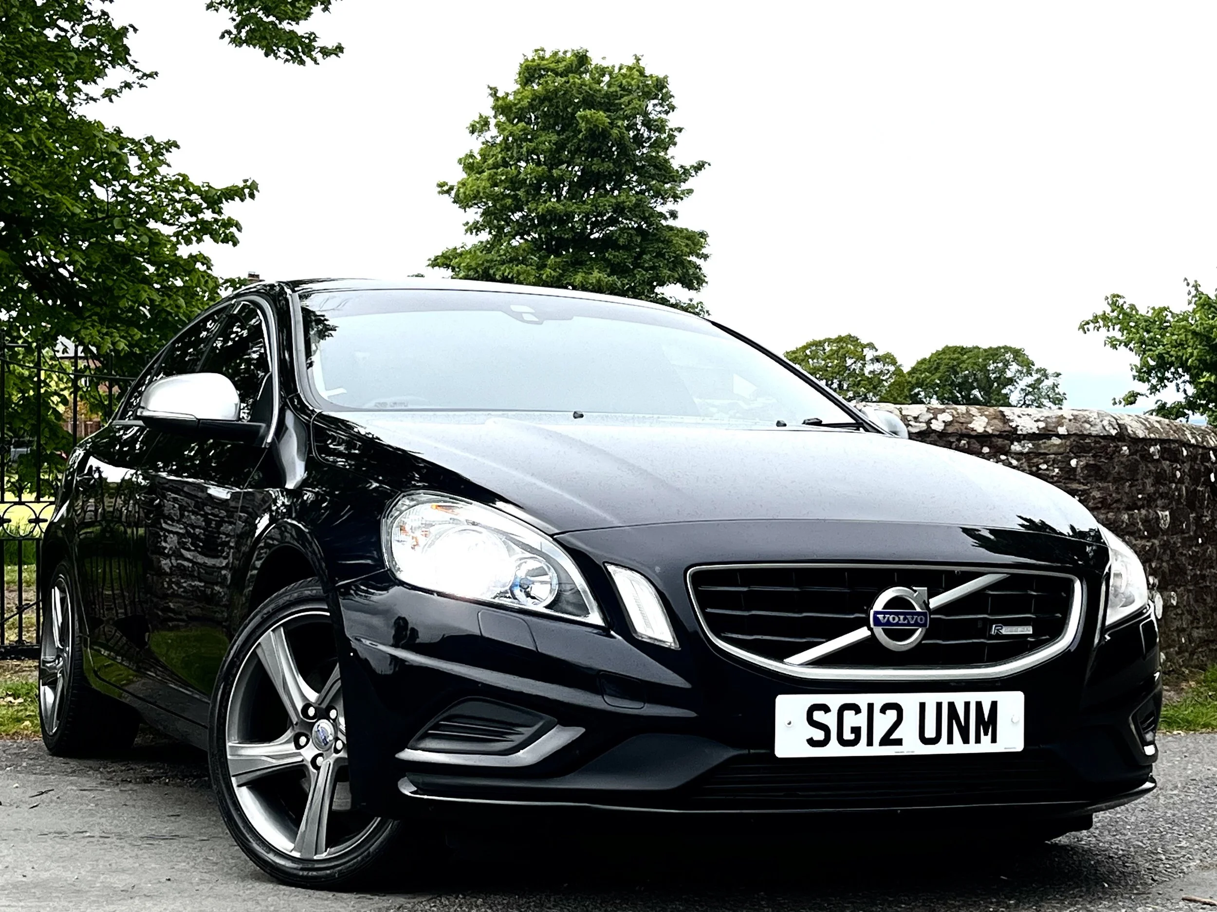 A black Volvo car parked outdoors, with a tree and cloudy sky in the background, and a white license plate with the text 'SG12 UNM'.