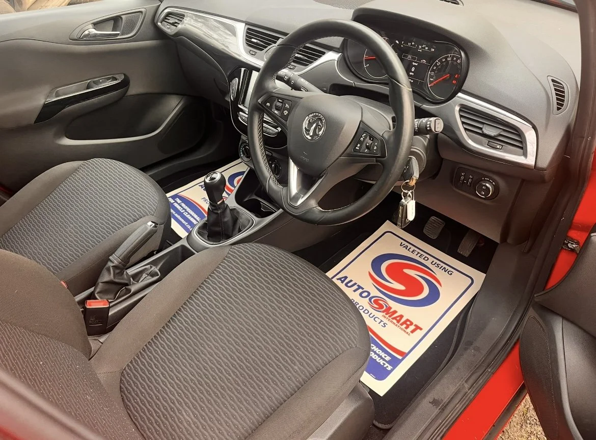 Interior view of a car showing the driver's seat, steering wheel, dashboard, gear shift, and floor mats with the logo and branding of 'AutoSmart'.