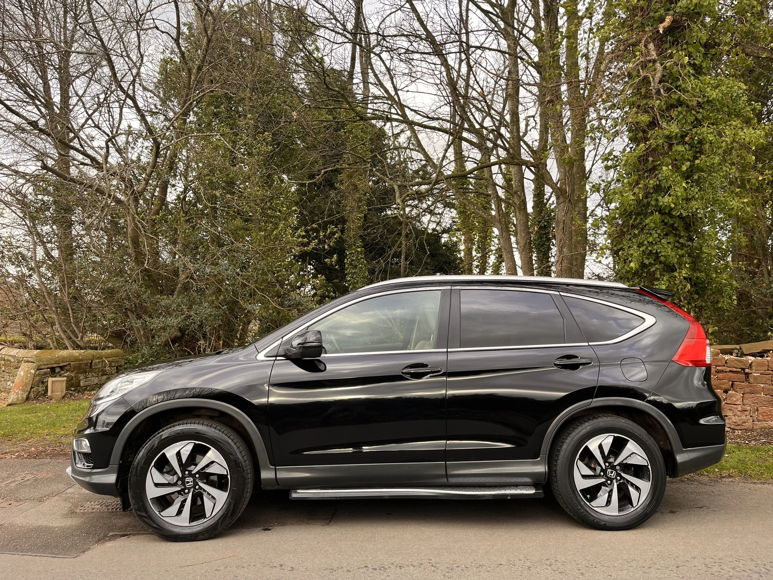 A black compact SUV parked on a quiet street with a background of leafless trees and a brick wall.
