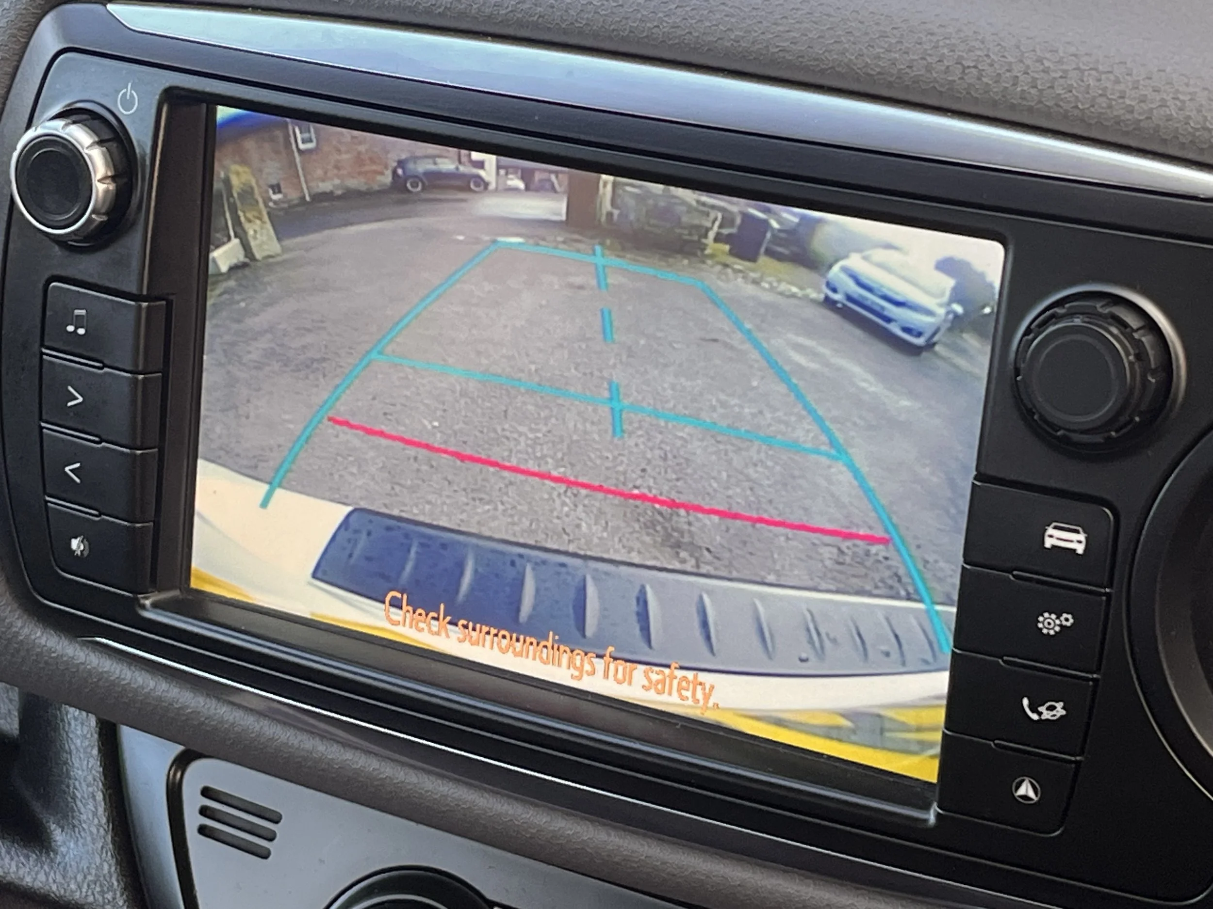 Car's dashboard camera display showing a rearview of a parking lot with cars and a brick building, alongside parking guidelines and a caution message.