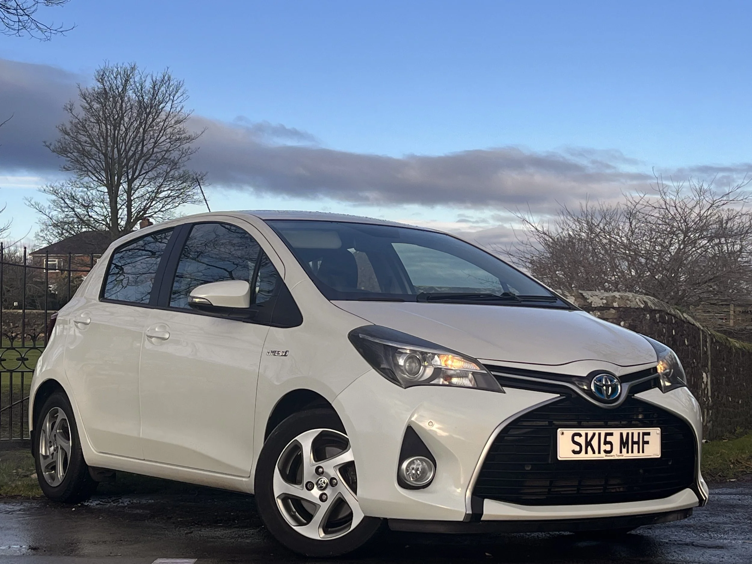 A white Toyota Yaris parked on a wet road with a fence, trees, and a partly cloudy sky in the background.
