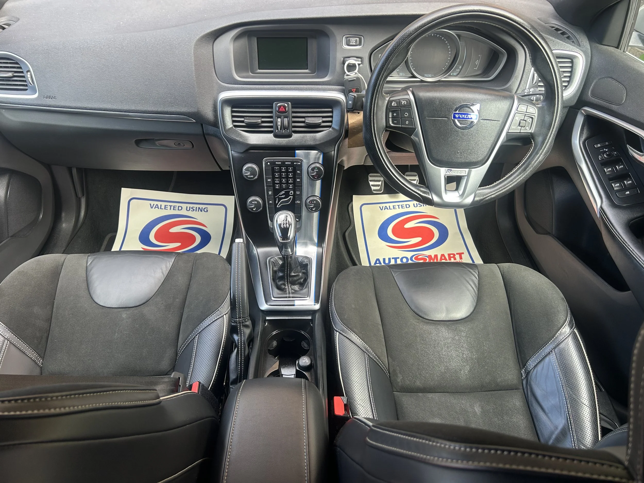 The interior of a car showing the dashboard, steering wheel, gear shift, and two front seats. The dashboard has a digital display, air vents, and control buttons. The car floor mats display the AUTO SMART logo and the words "Valeted Using".