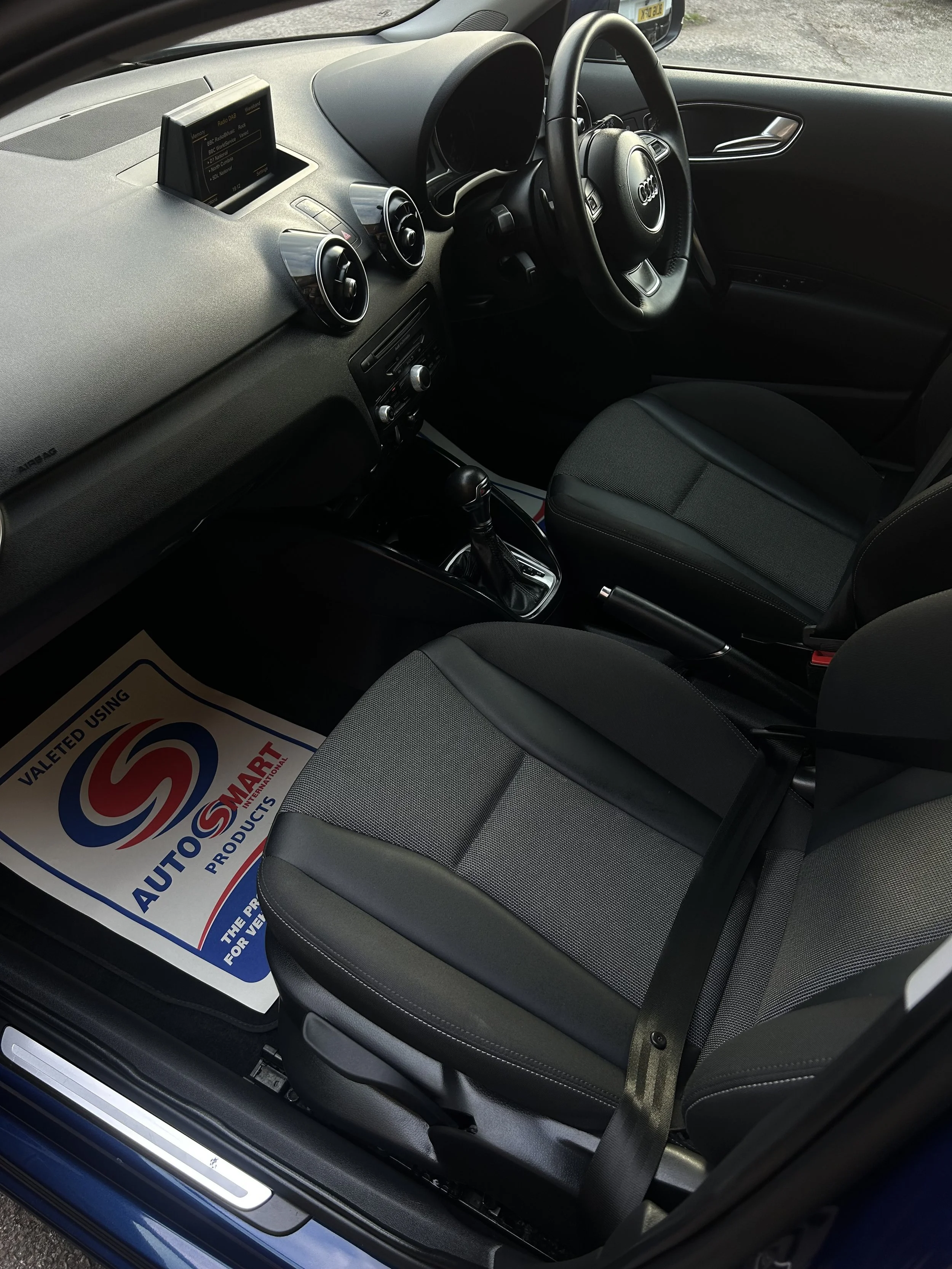 Interior of a car showing the driver's seat, passenger seat, steering wheel with Audi logo, dashboard, and gear shift, with a paper mat on the passenger side floor.