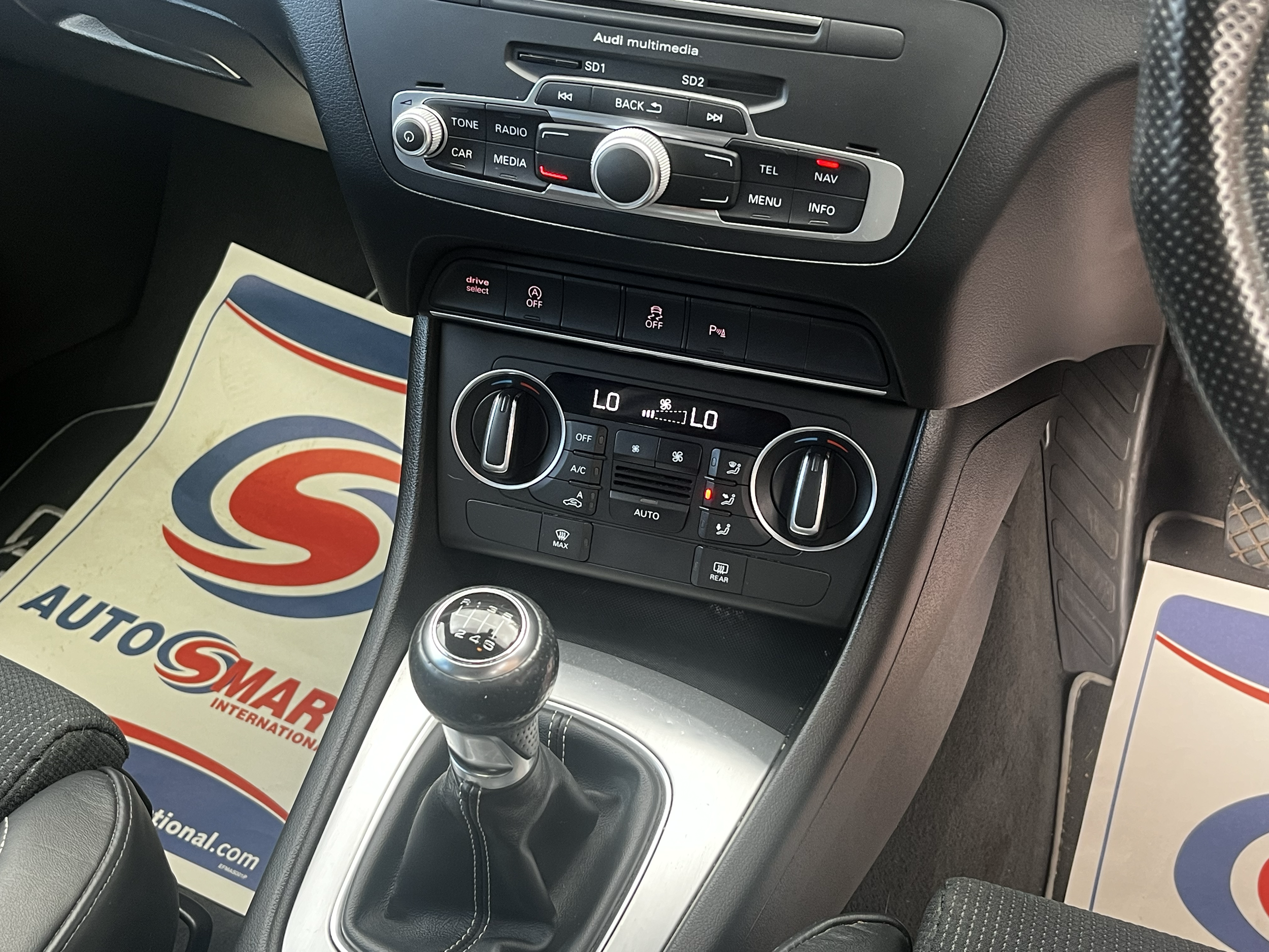 Center console and dashboard of a car with multimedia controls, climate controls, and a manual gear shift. A paper mat with red and blue logos is on the car floor.
