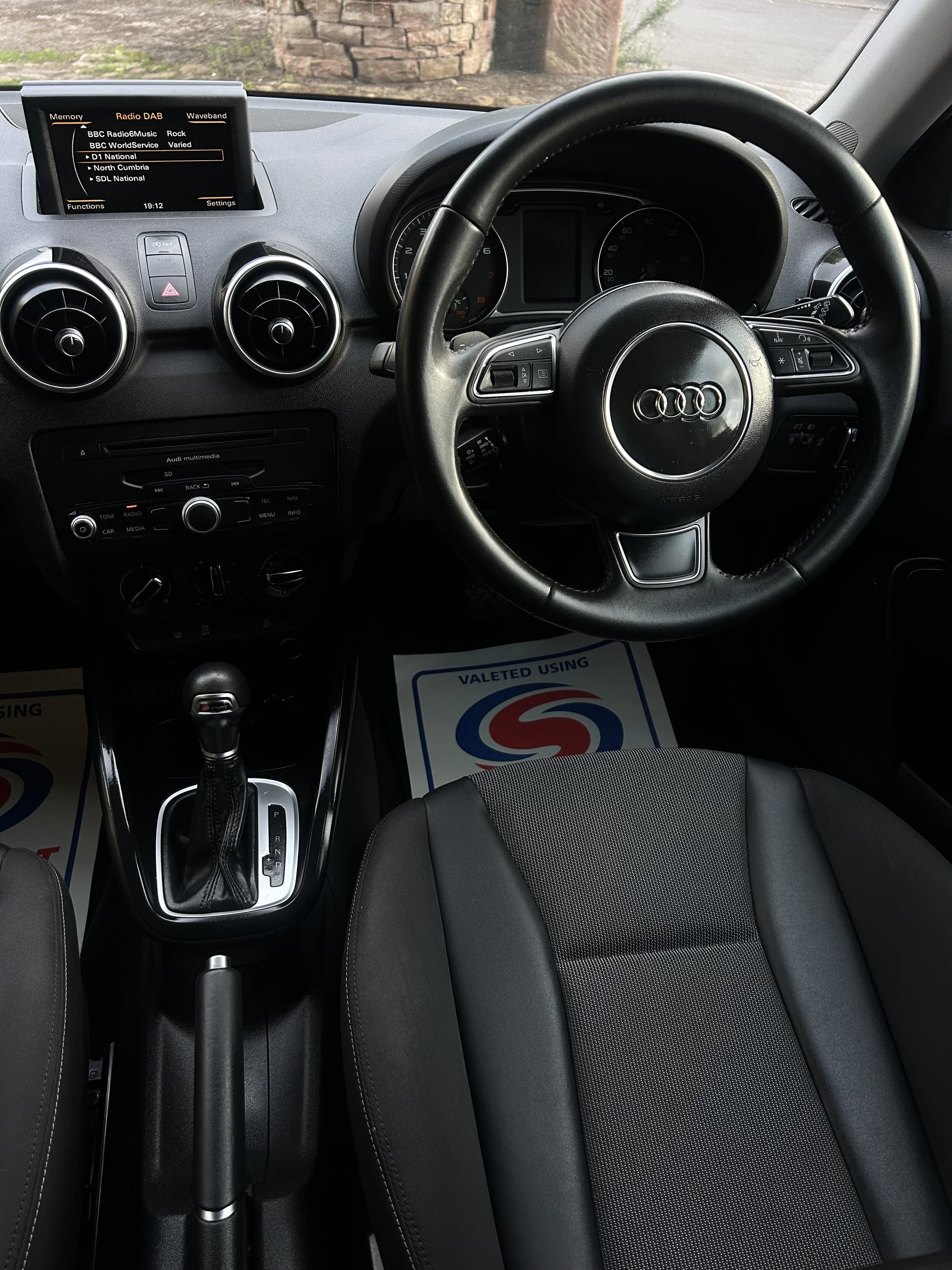 Interior of an Audi car showing the steering wheel with the Audi logo, dashboard, gear shift, and a partial view of the driver's seat.