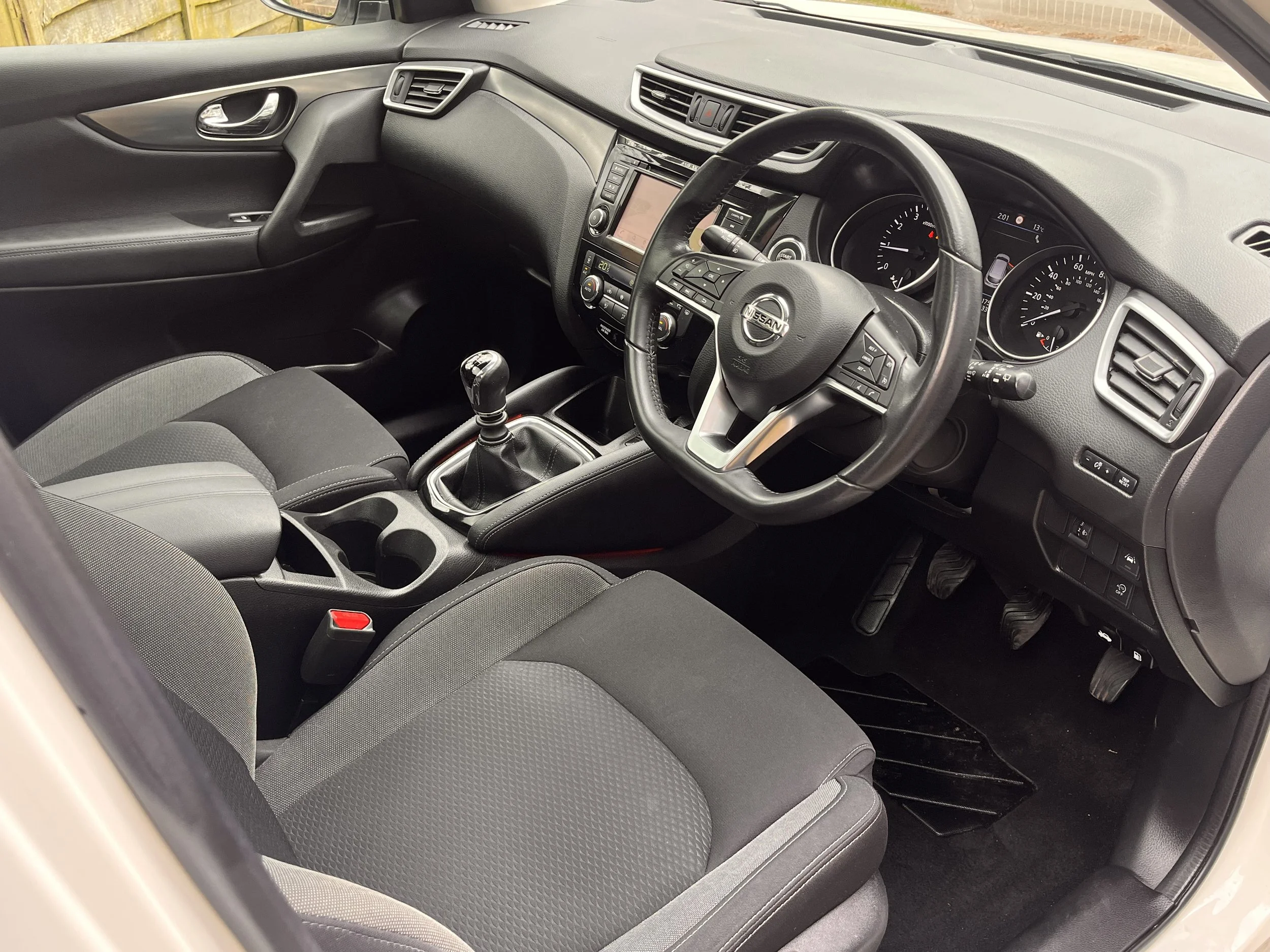 Interior of a Nissan vehicle showing the steering wheel, gear shift, dashboard with digital display, and front seats with fabric upholstery.