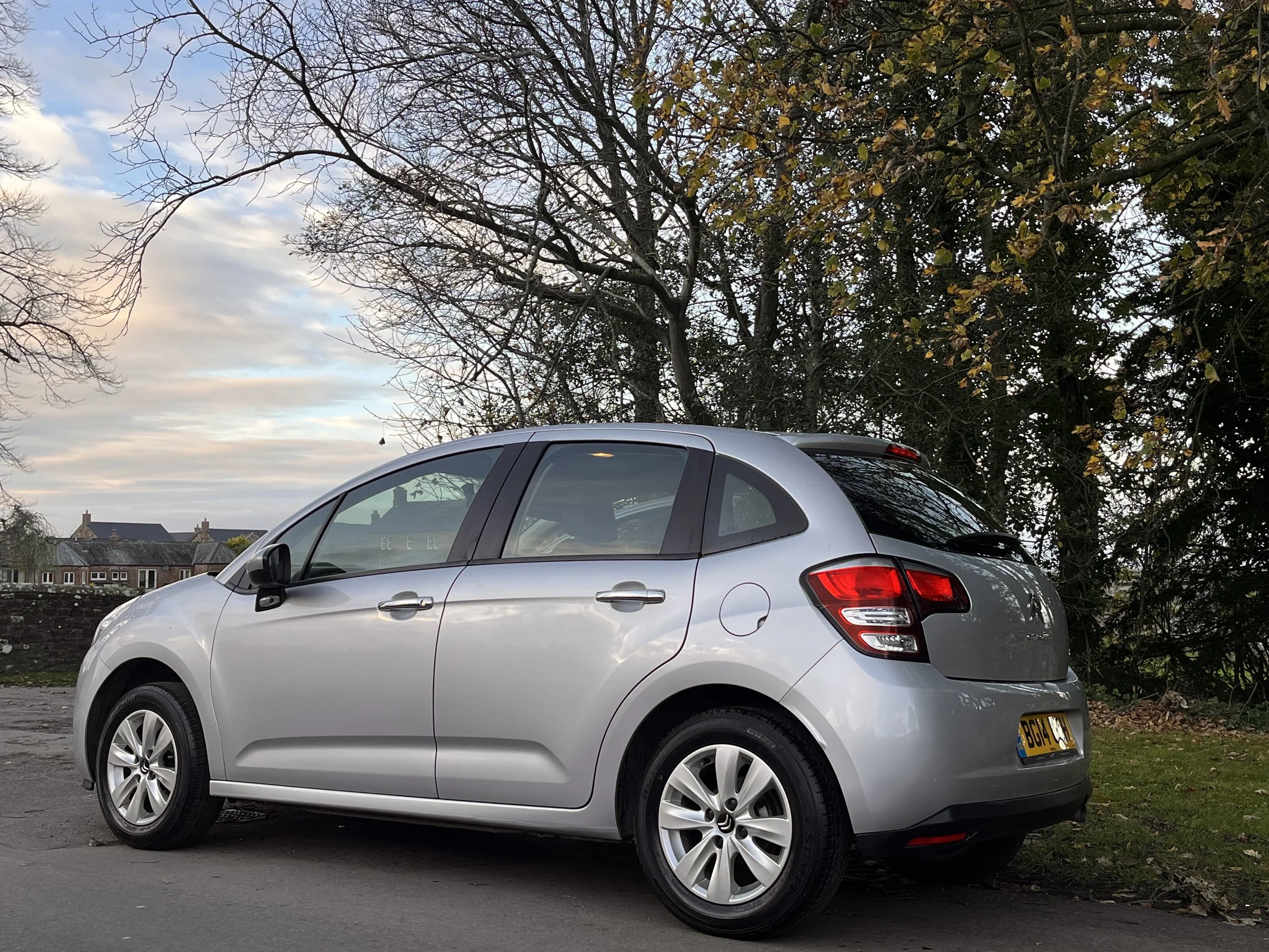 A silver compact hatchback car parked outdoors on a paved surface, with trees and houses in the background during the daytime.