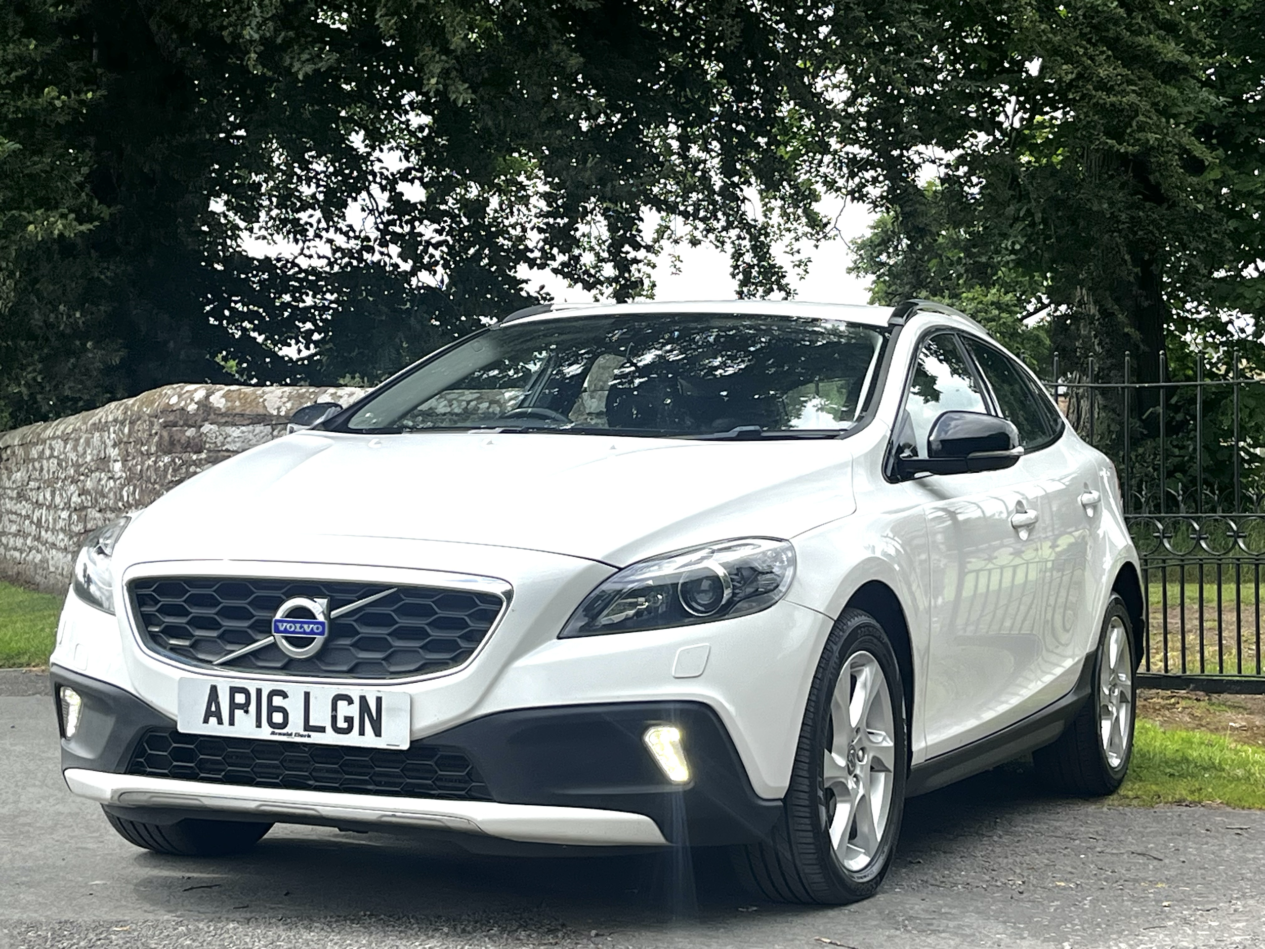 A white Volvo car is parked on a street next to a stone wall. The car has a UK license plate, and the background features large leafy trees and a metal fence.