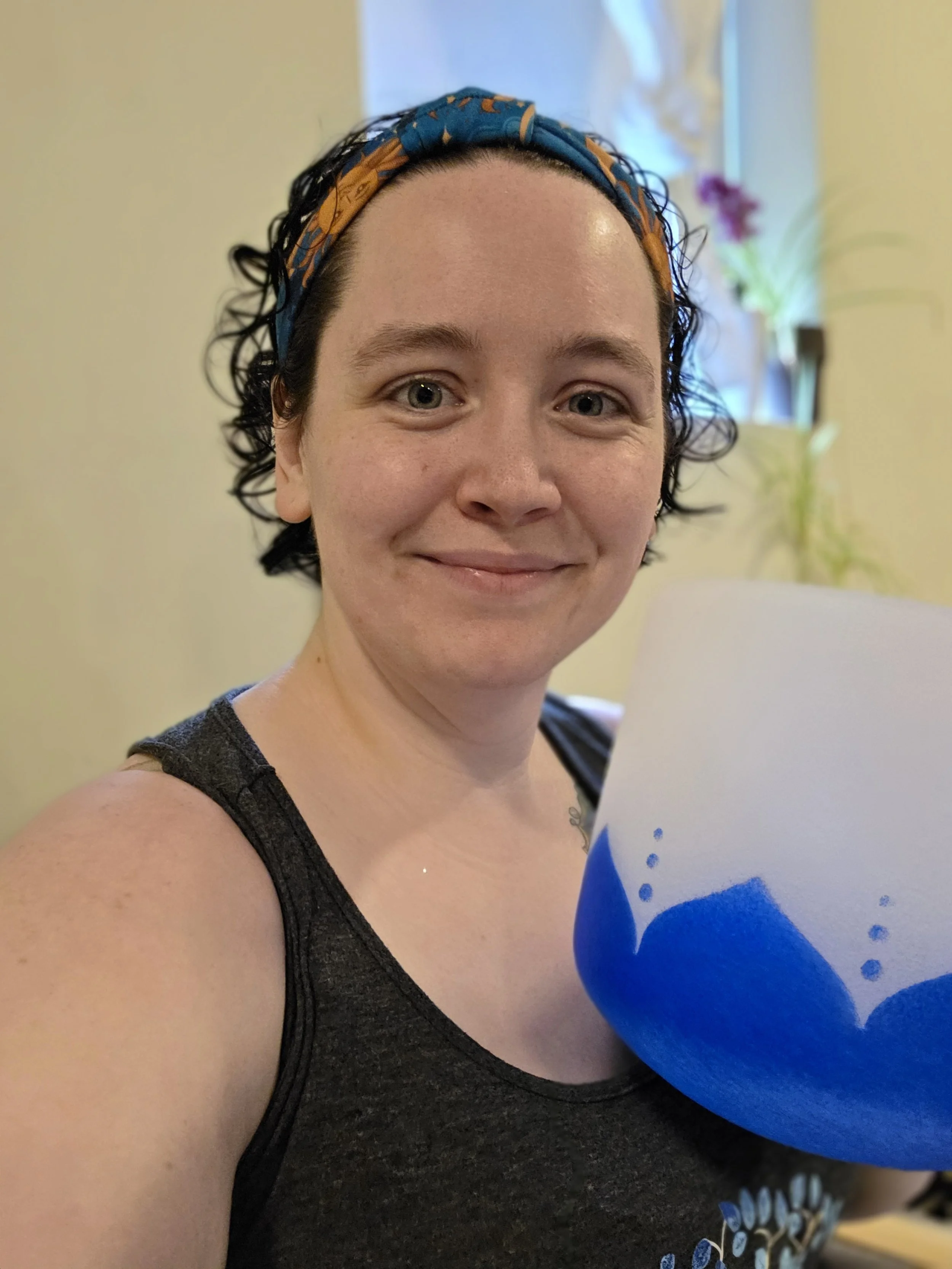 A woman with short, curly dark hair wearing a blue and orange headband taking a selfie inside a room with a yellow wall and plants in the background. She is smiling and holding a large bowl with blue and white patterns.