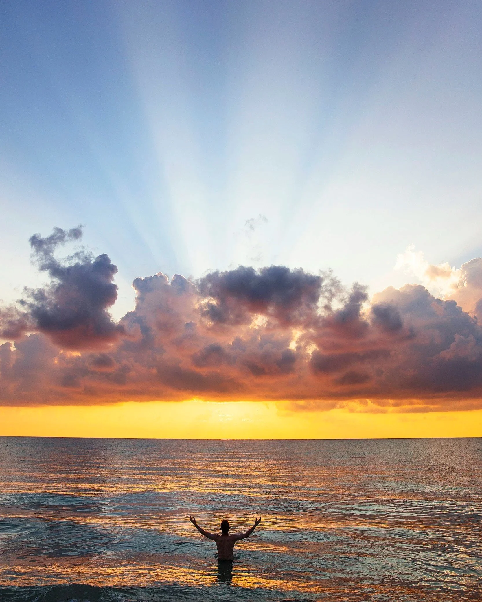 Person standing in ocean water at sunset with arms raised, colorful sky with clouds and sun rays above the faith integration into therapy section.