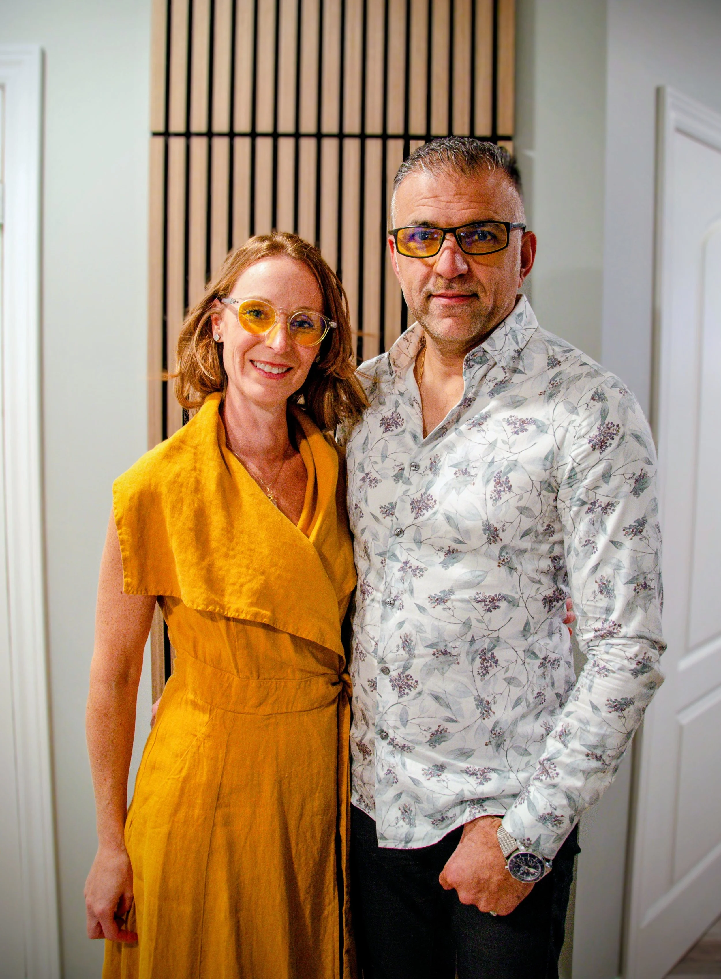 A woman and a man standing close together indoors, both wearing yellow tinted glasses. The woman has short red hair, is smiling, and is dressed in a yellow dress, while the man has short gray hair, is wearing glasses, and dressed in a white shirt with a floral pattern, and black pants.