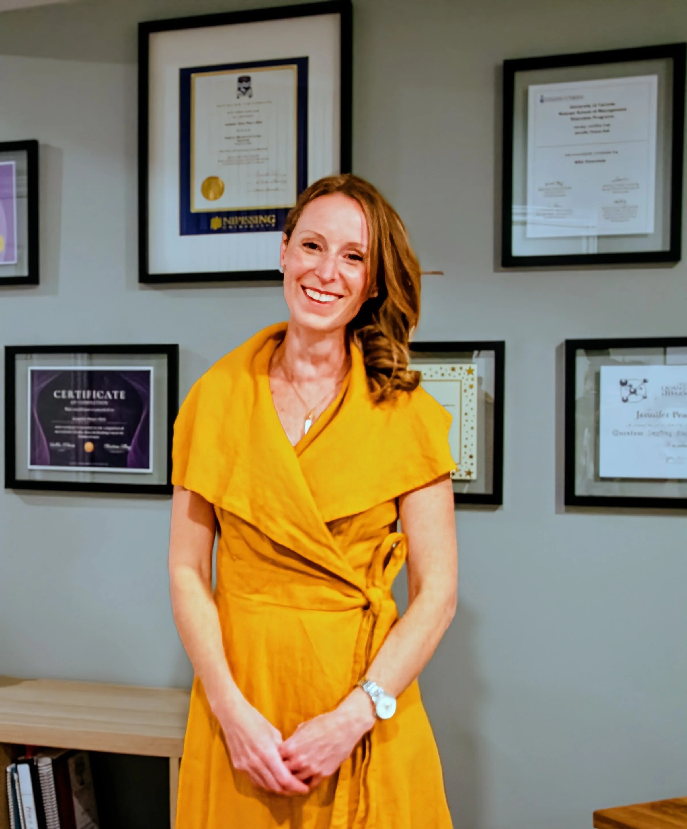 Smiling woman in a yellow dress standing indoors in front of framed certificates or diplomas on a wall.
