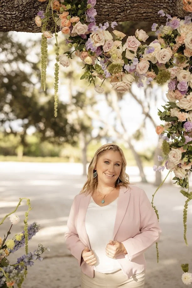 A woman standing outdoors under a floral arch with pink, purple, and white flowers, smiling and looking at the camera.