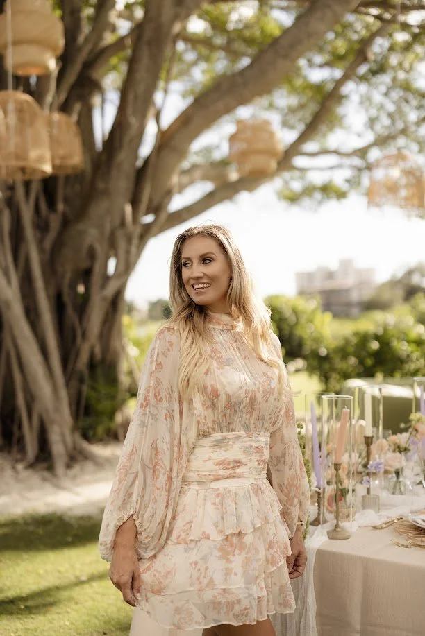 A woman with long blonde hair, wearing a pastel-colored floral dress, smiling outdoors during daytime, with a table decorated with candles and flowers nearby and a large tree in the background.