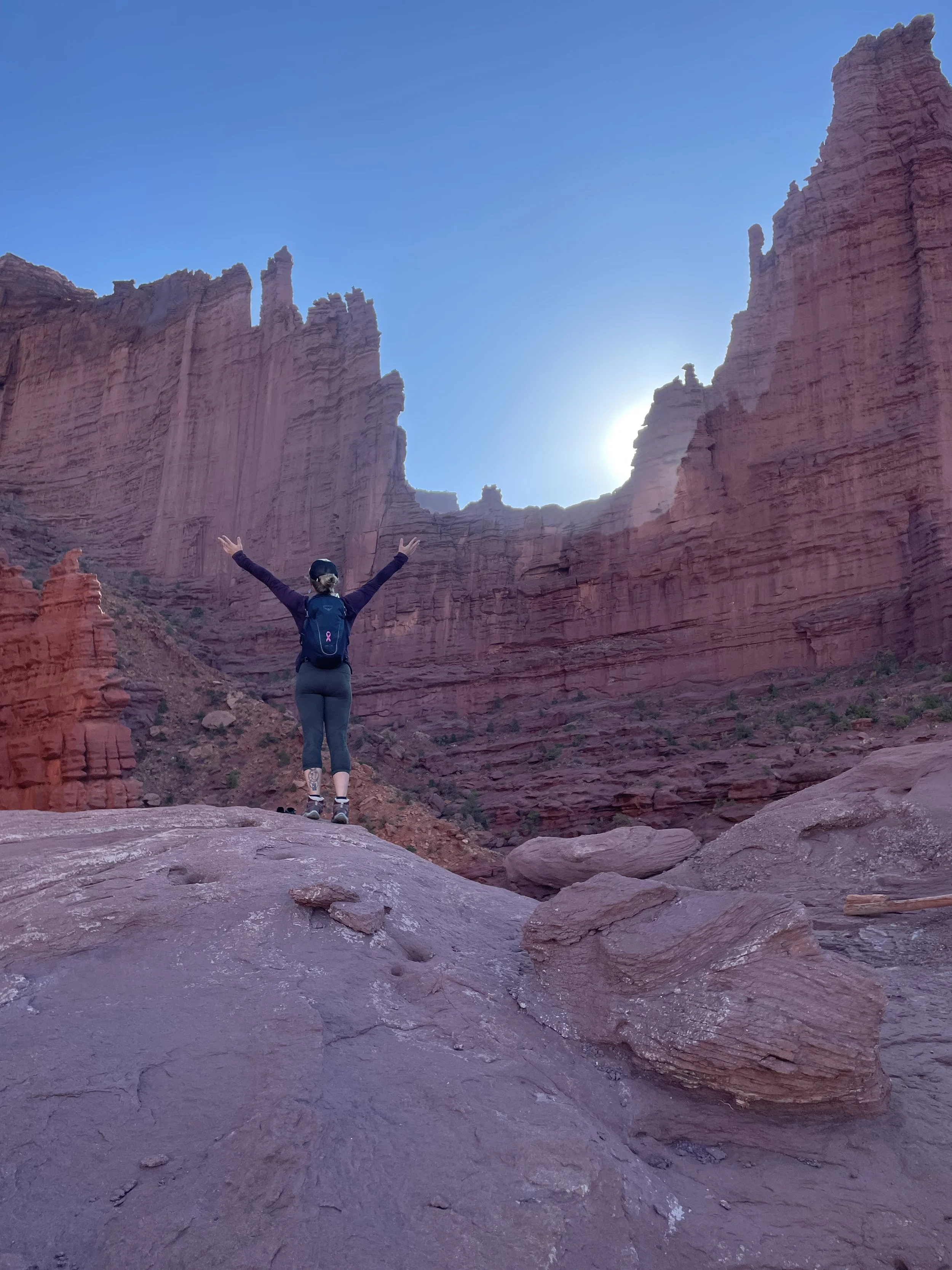 Person standing on rocks with arms raised in front of towering red rock cliffs at a canyon, with the sun shining behind the cliffs and a clear blue sky.