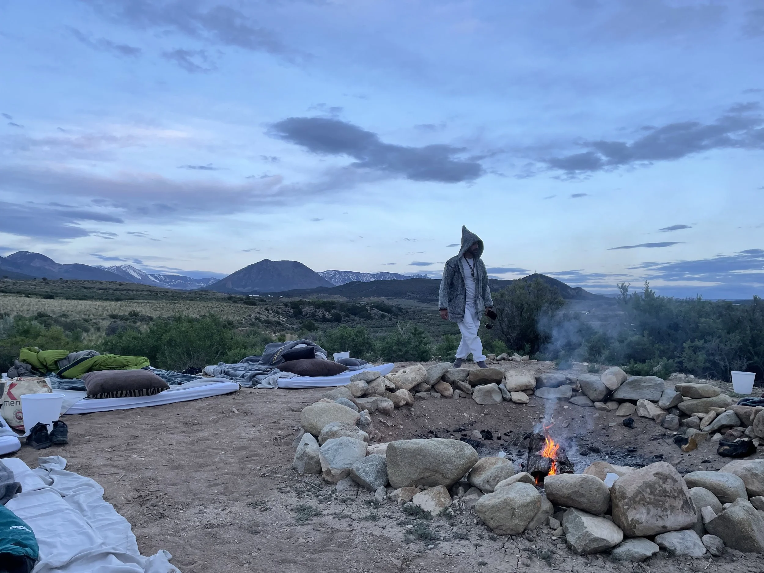 Person in hooded jacket walking around a campfire with sleeping bags and backpacks on the ground, mountains in the background, during dusk.